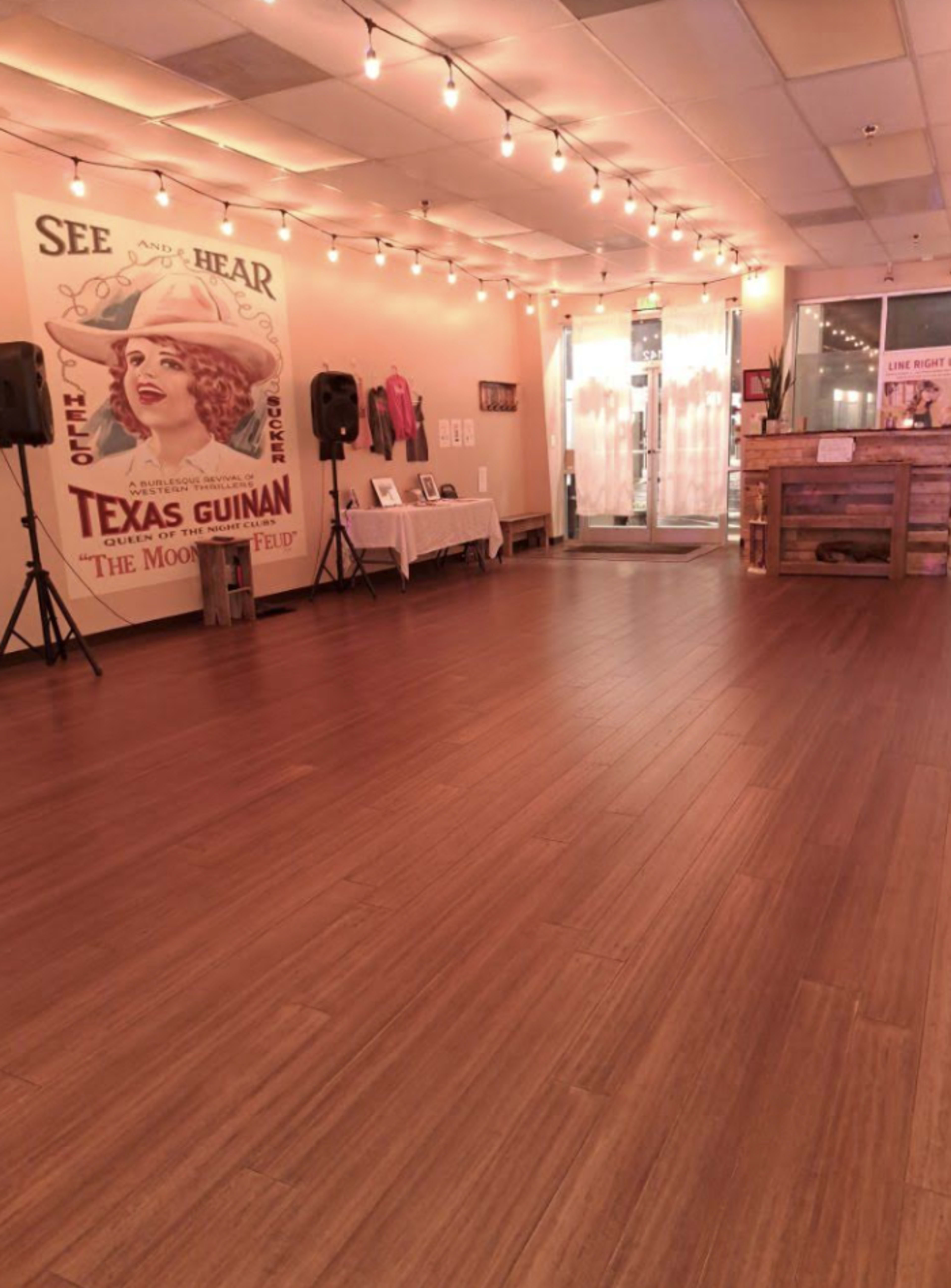 An interior space with wooden flooring, string lights overhead, a vintage poster of Texas Guinan on the wall, and a wooden reception counter near the entrance.