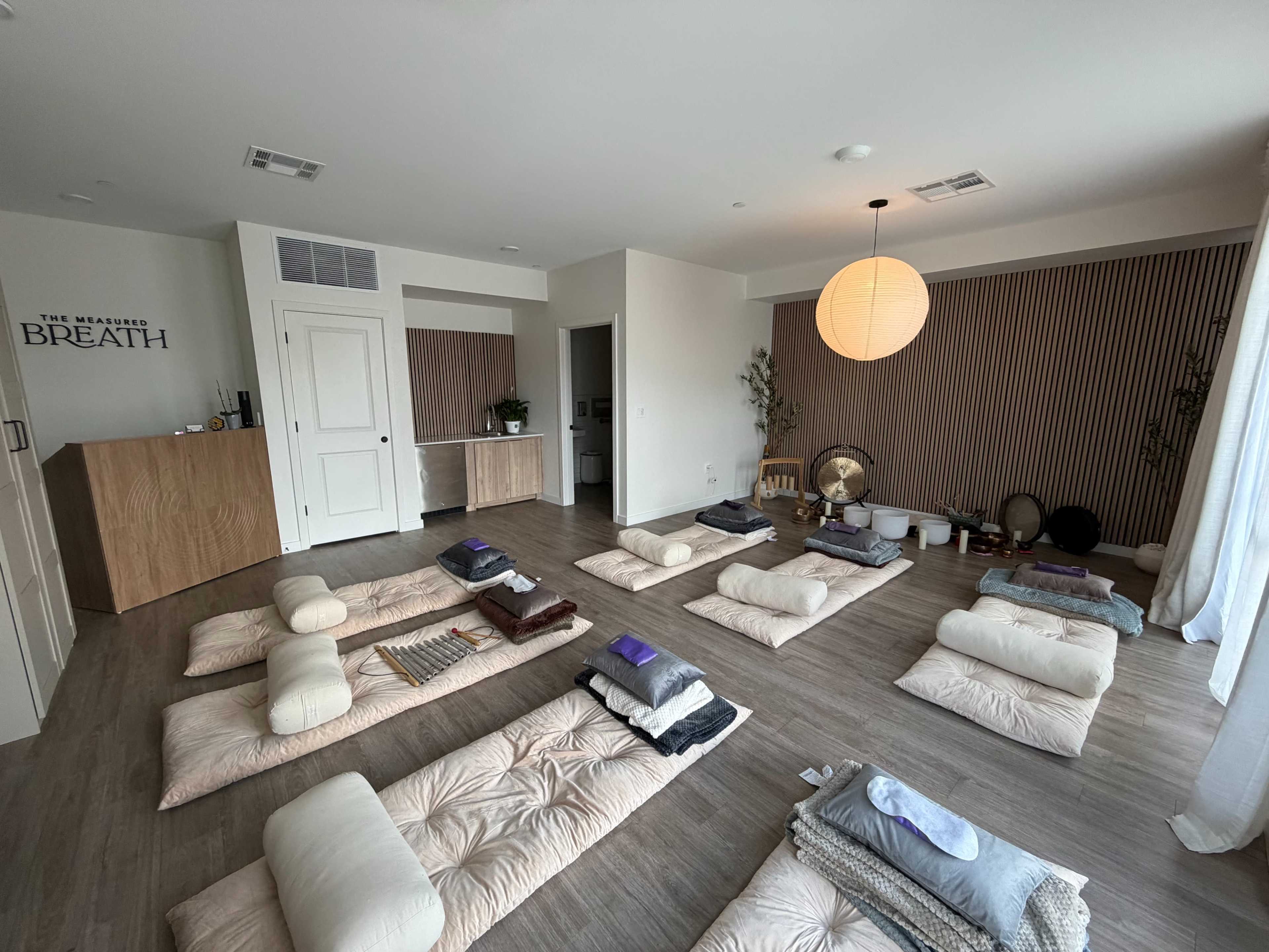 The image shows a tranquil wellness studio with several mats and cushions arranged on the floor, a warm light fixture, and a minimalist decor.