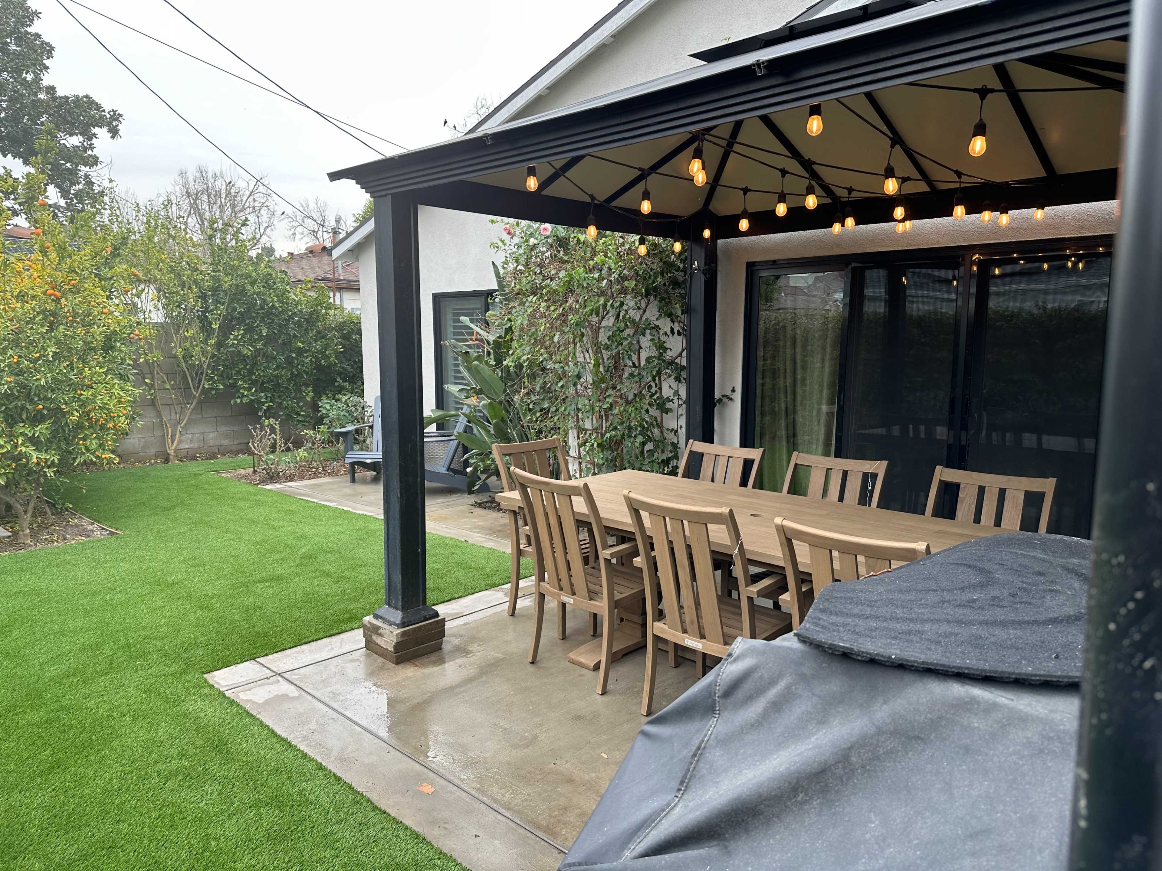 A patio with a large wooden dining table and chairs under a black canopy, surrounded by green grass and plants.