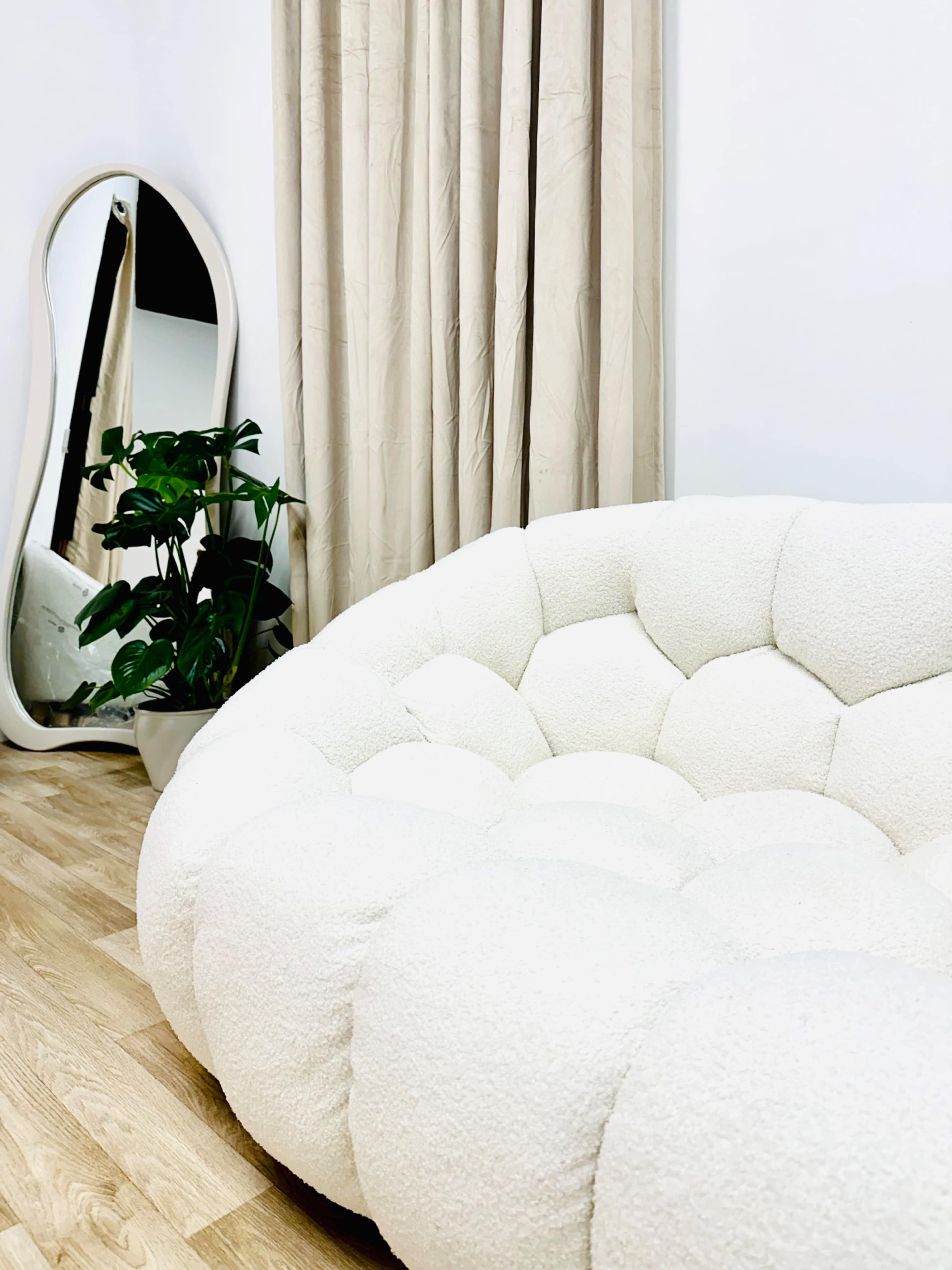 The image shows a plush, round white couch positioned next to a tall plant and a full-length mirror, with light-colored curtains in the background.
