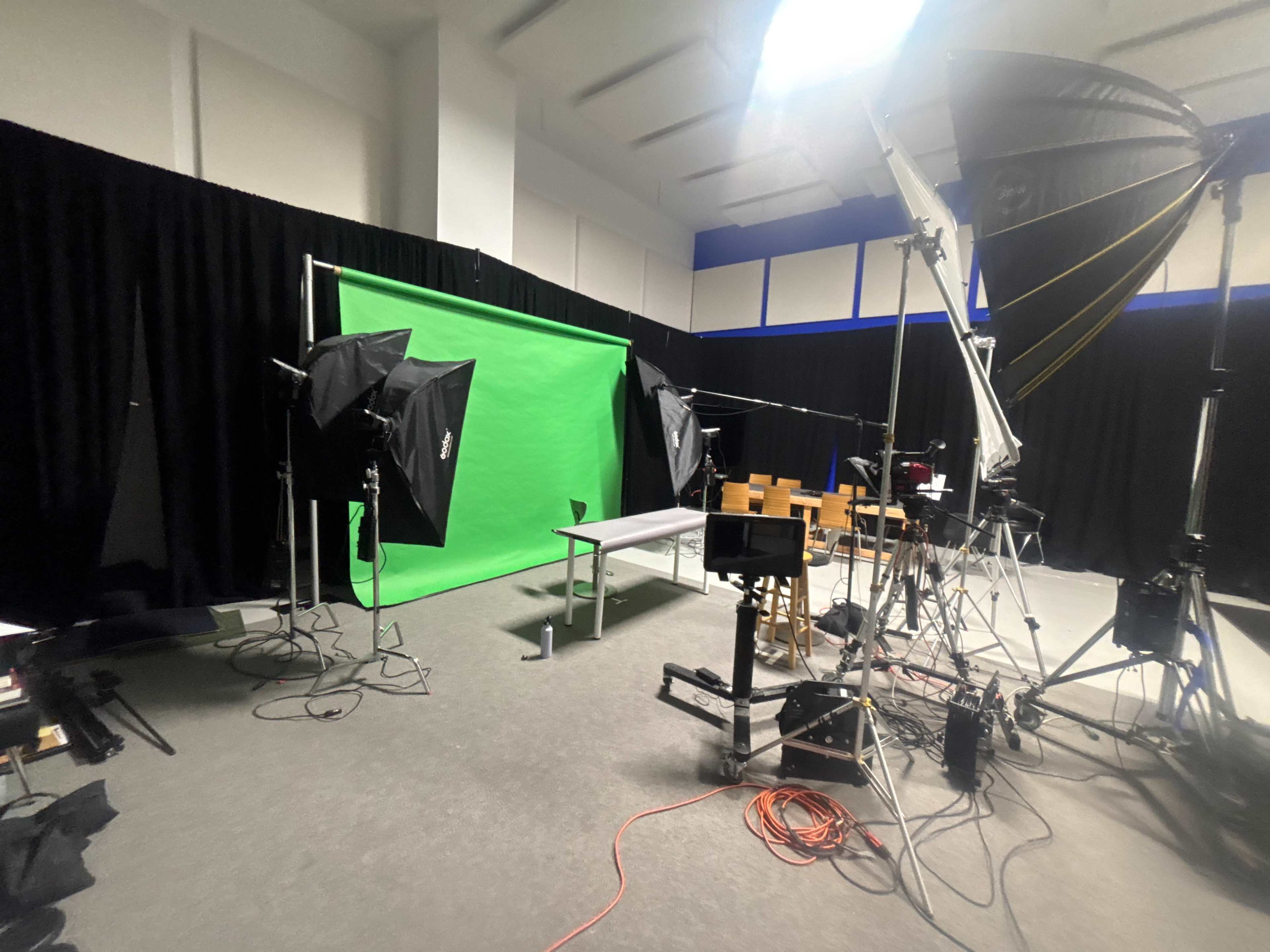 The image shows a studio setup with a green screen, lighting equipment, and cameras arranged around a table and chairs.