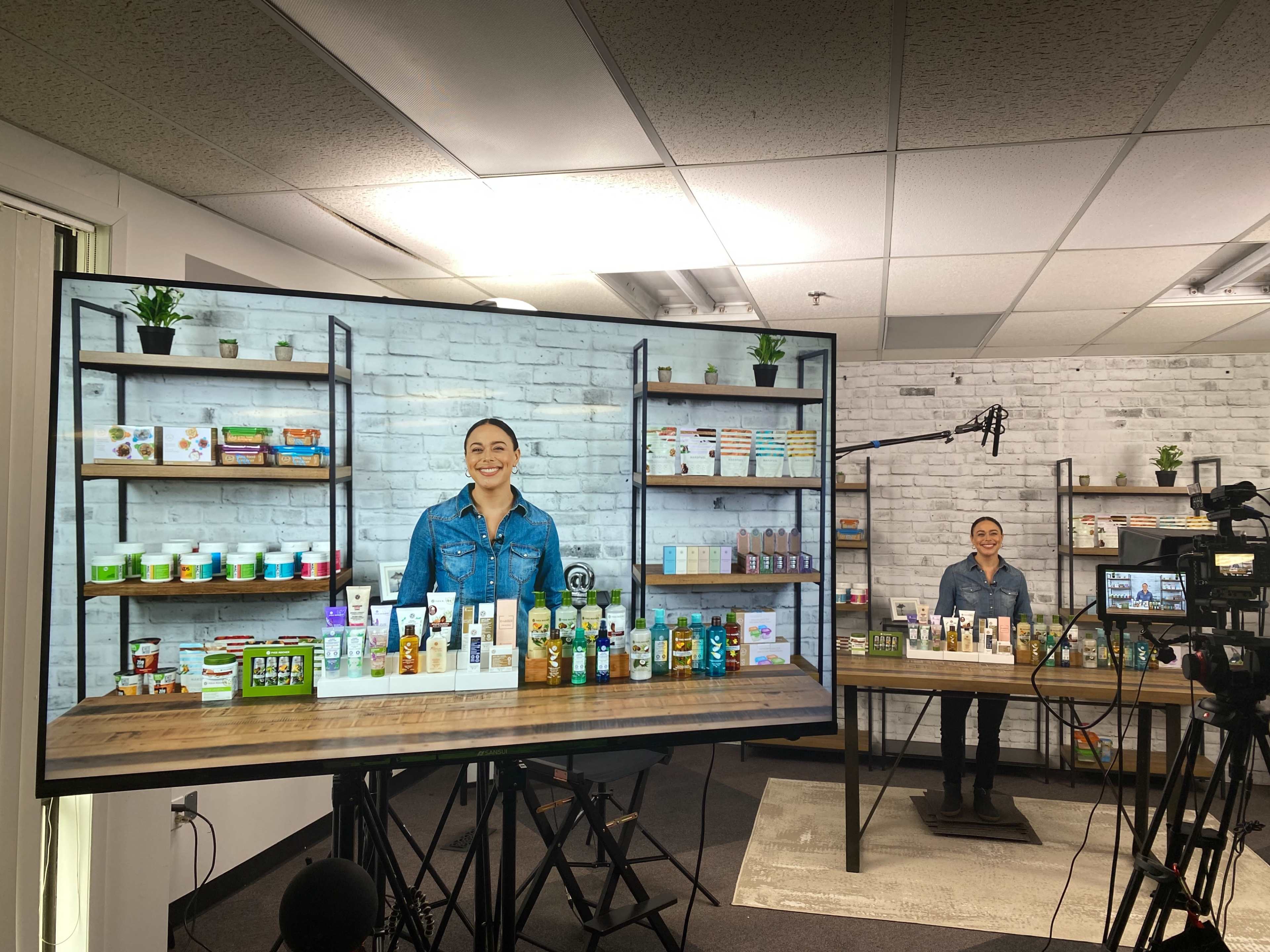 A woman stands behind a table displaying various products, with a large screen showing her image and a backdrop of shelves filled with additional items.