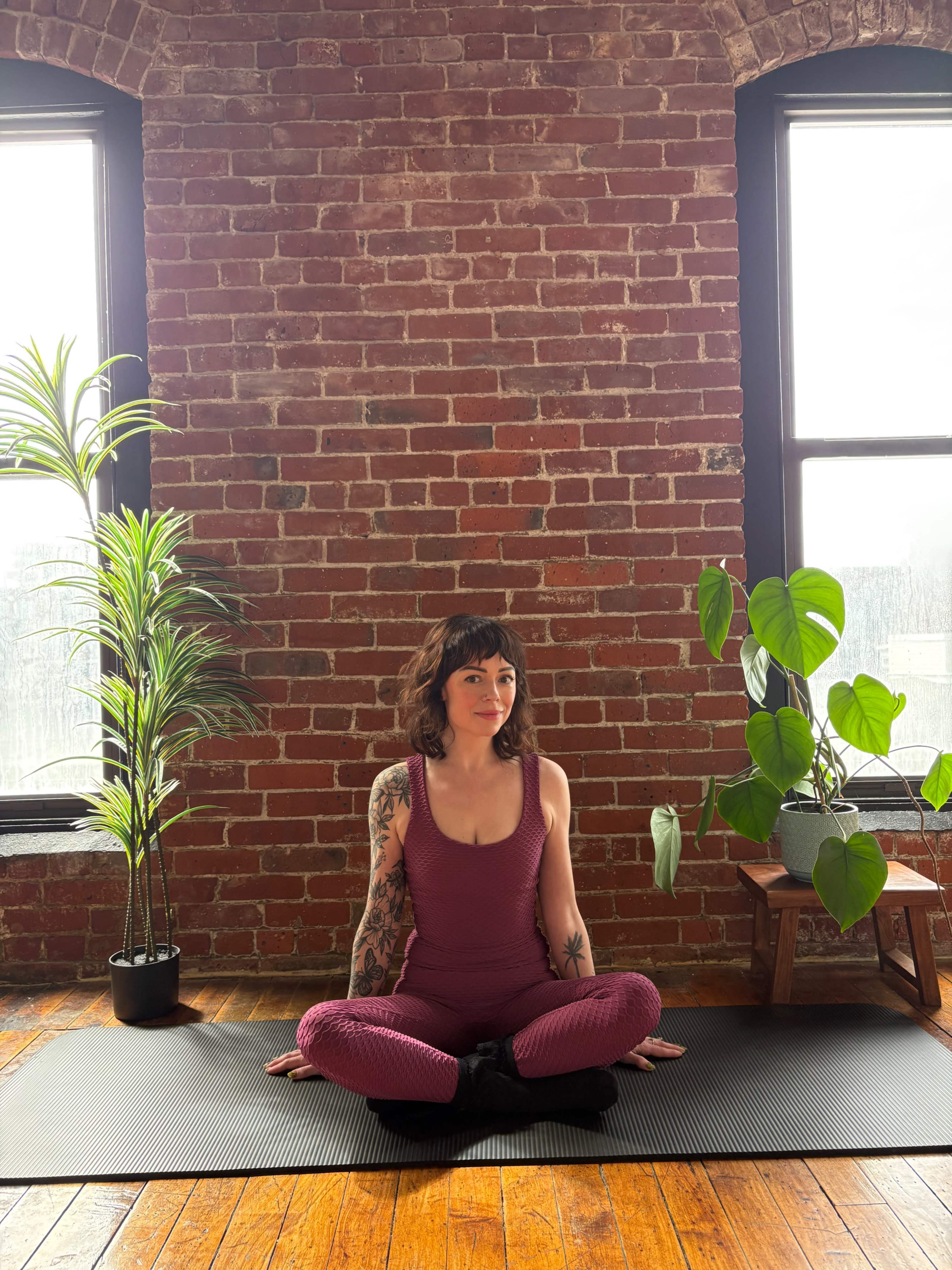A person sits cross-legged on a yoga mat in front of a brick wall, surrounded by indoor plants with large leaves.