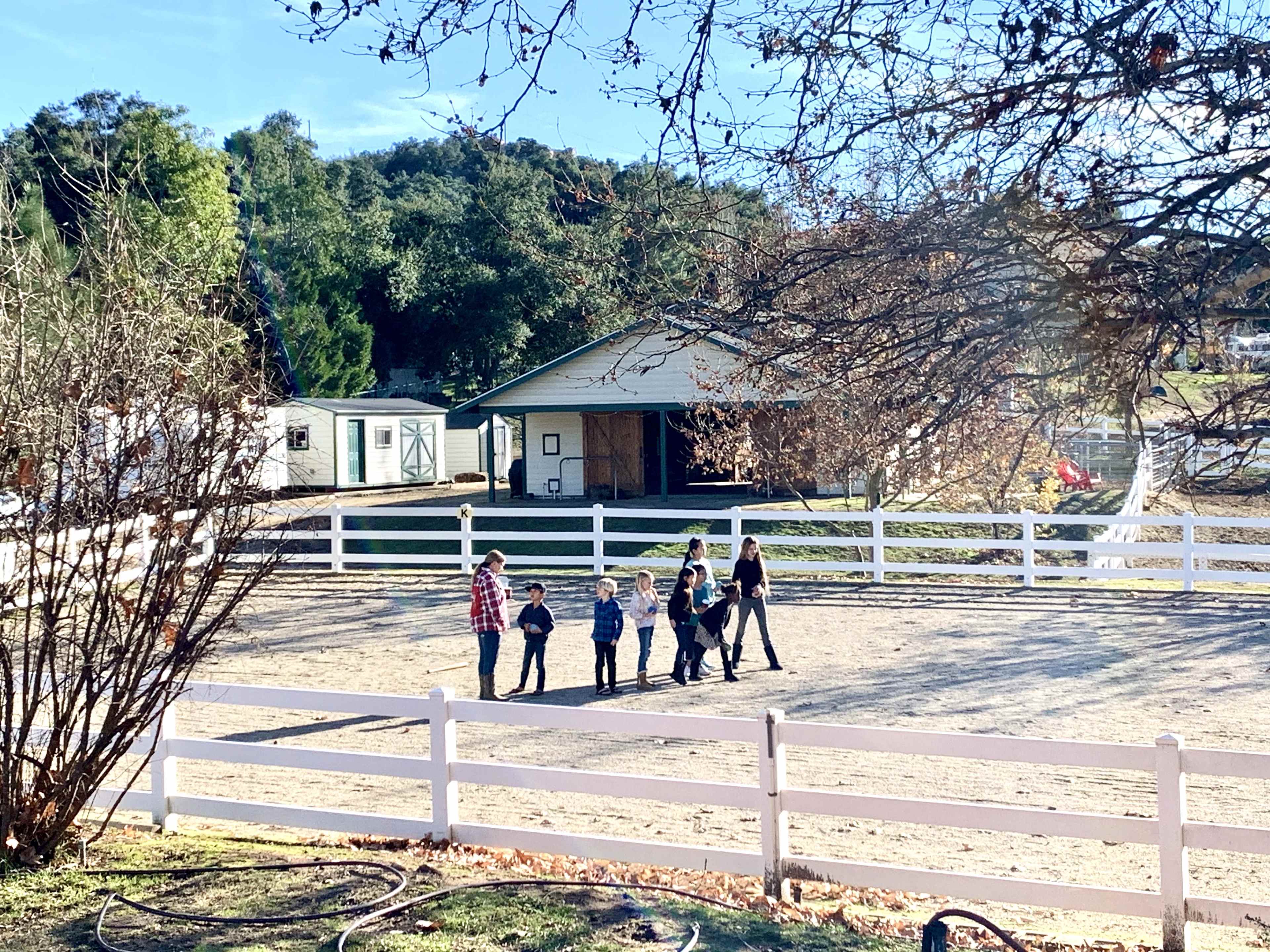 A group of children and adults walks along a sandy area near a white wooden fence and a building in the background.