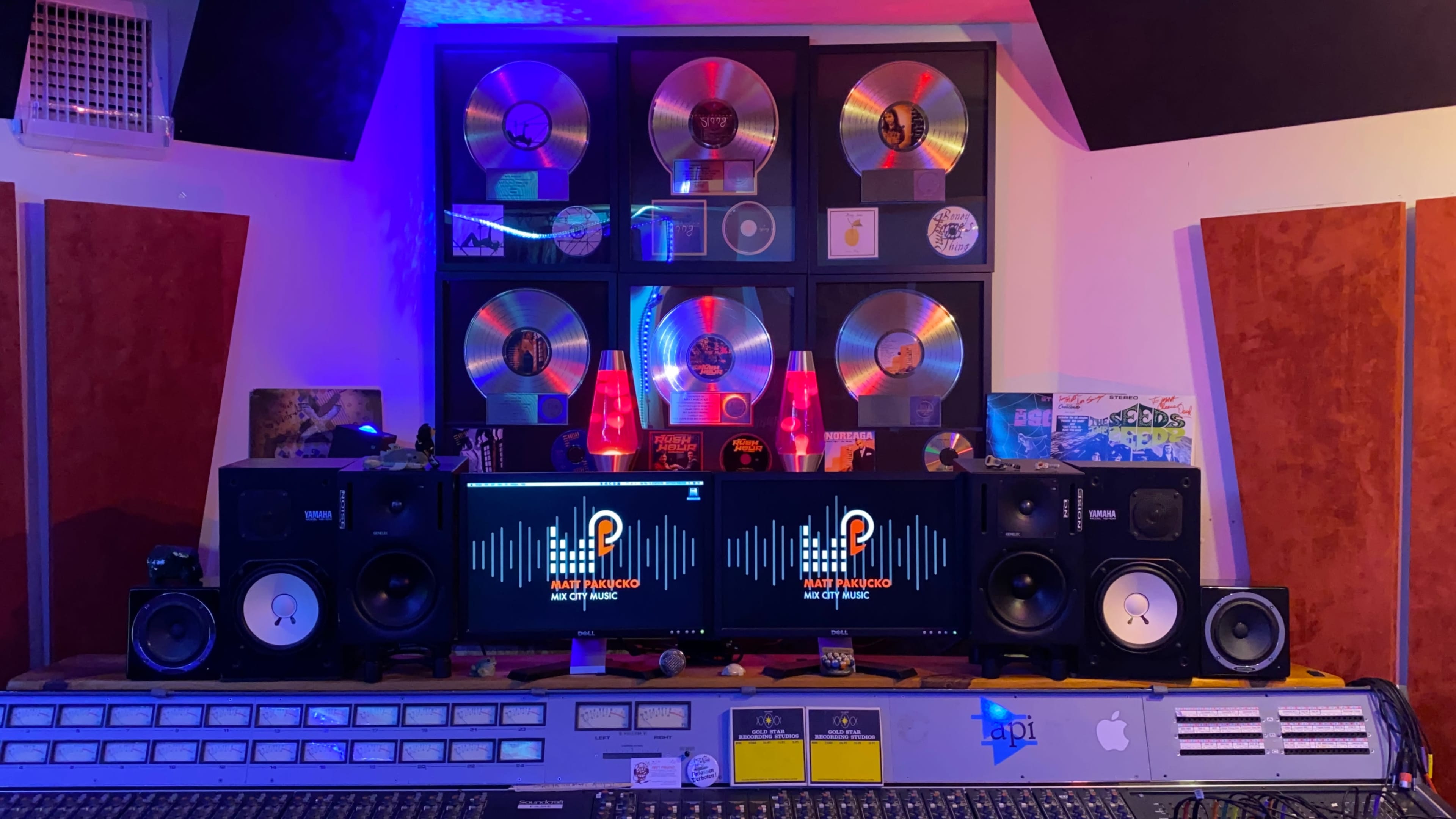 The image features a music studio with a decorative wall of gold and platinum records above a sound mixing console and two computer monitors displaying audio waveforms.