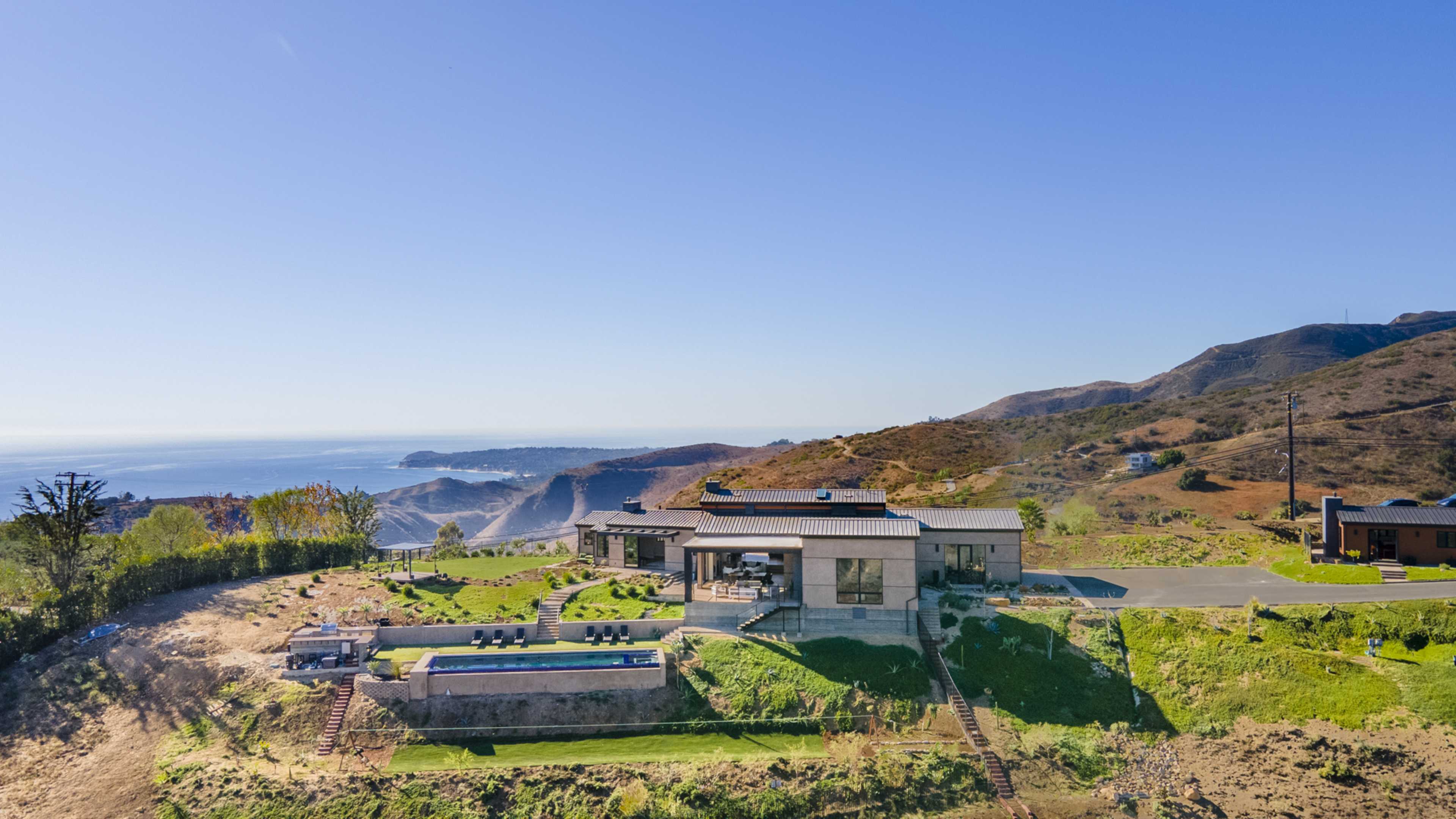 A modern house with a landscaped yard overlooks a coastal view and rolling hills.
