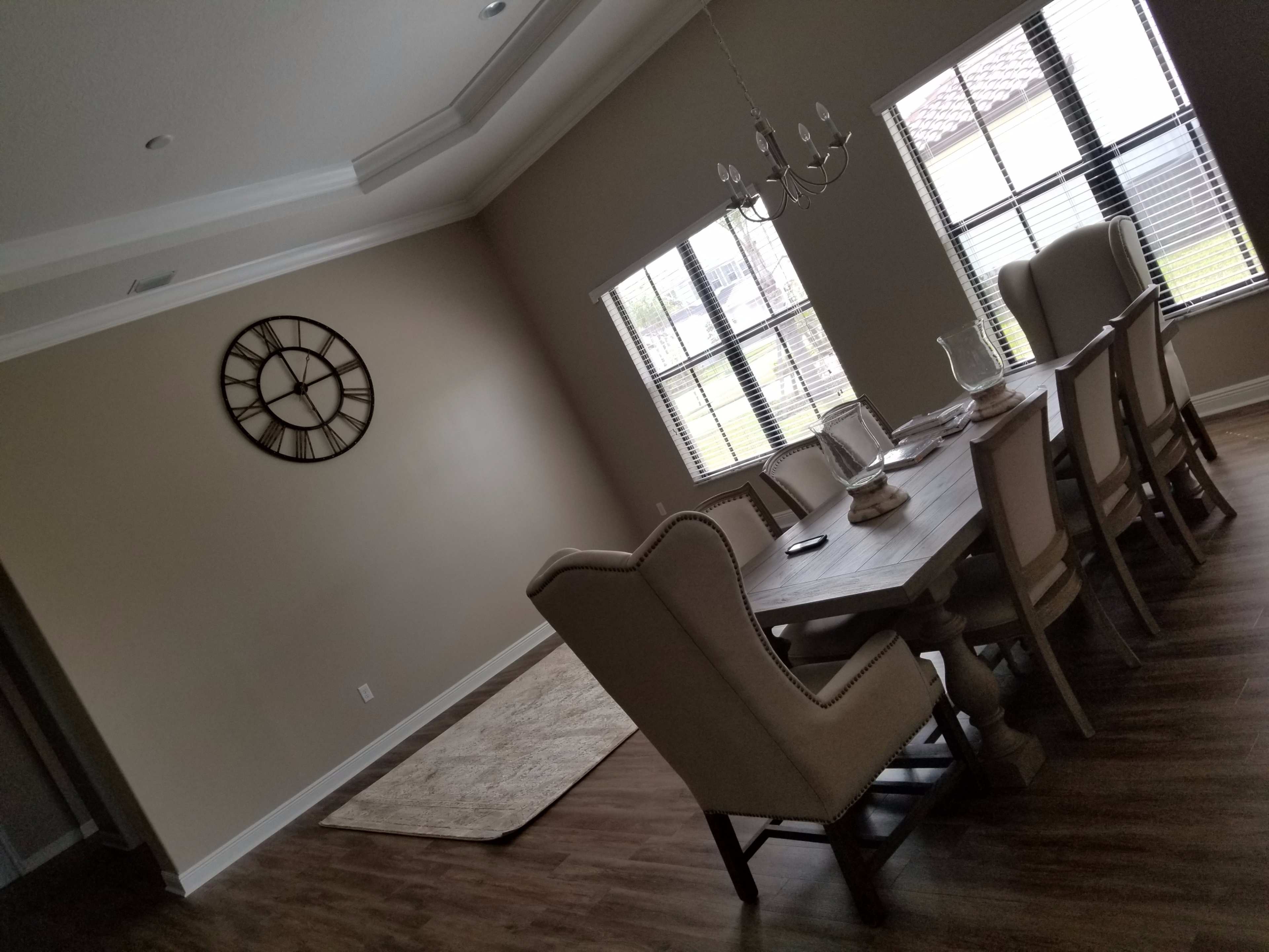 A dining room features a large table surrounded by upholstered chairs, with a clock on the wall and windows allowing natural light.