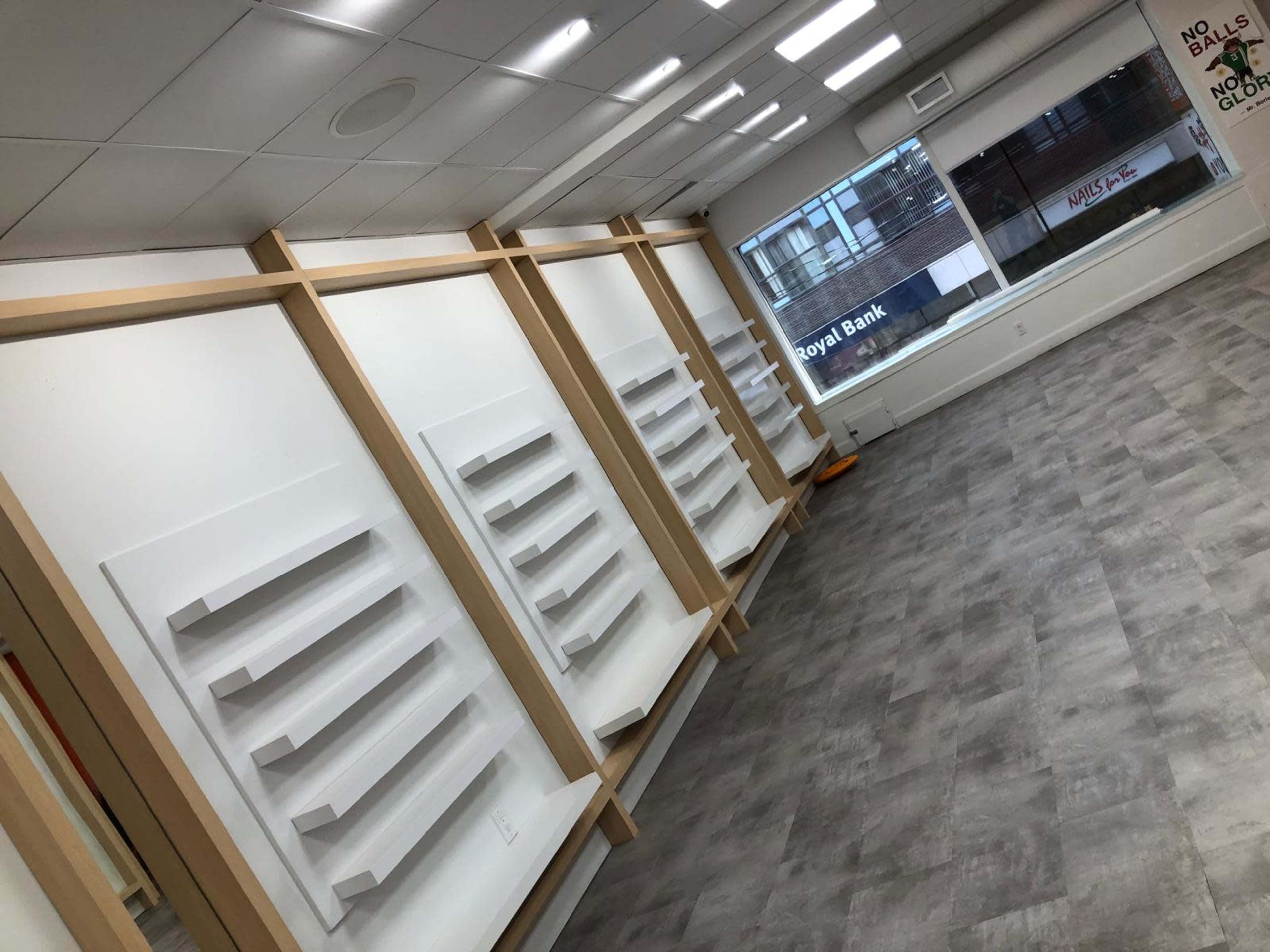 The image shows a bright, empty room with several white shelves mounted on wooden frames along the walls, and large windows allowing natural light to enter.