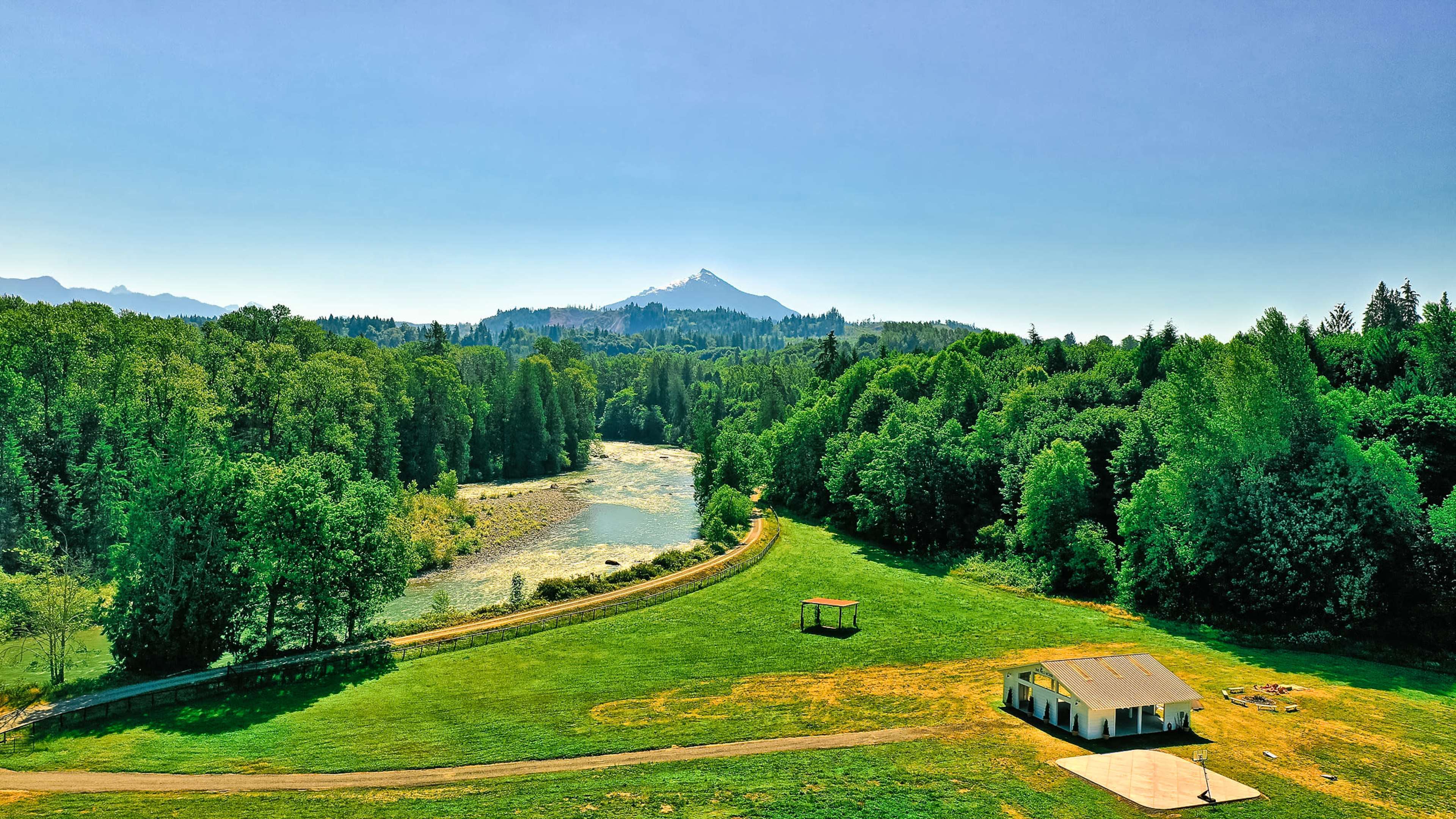 A lush green landscape with a winding river, bordered by trees, and a small building in the foreground with a mountain in the background.
