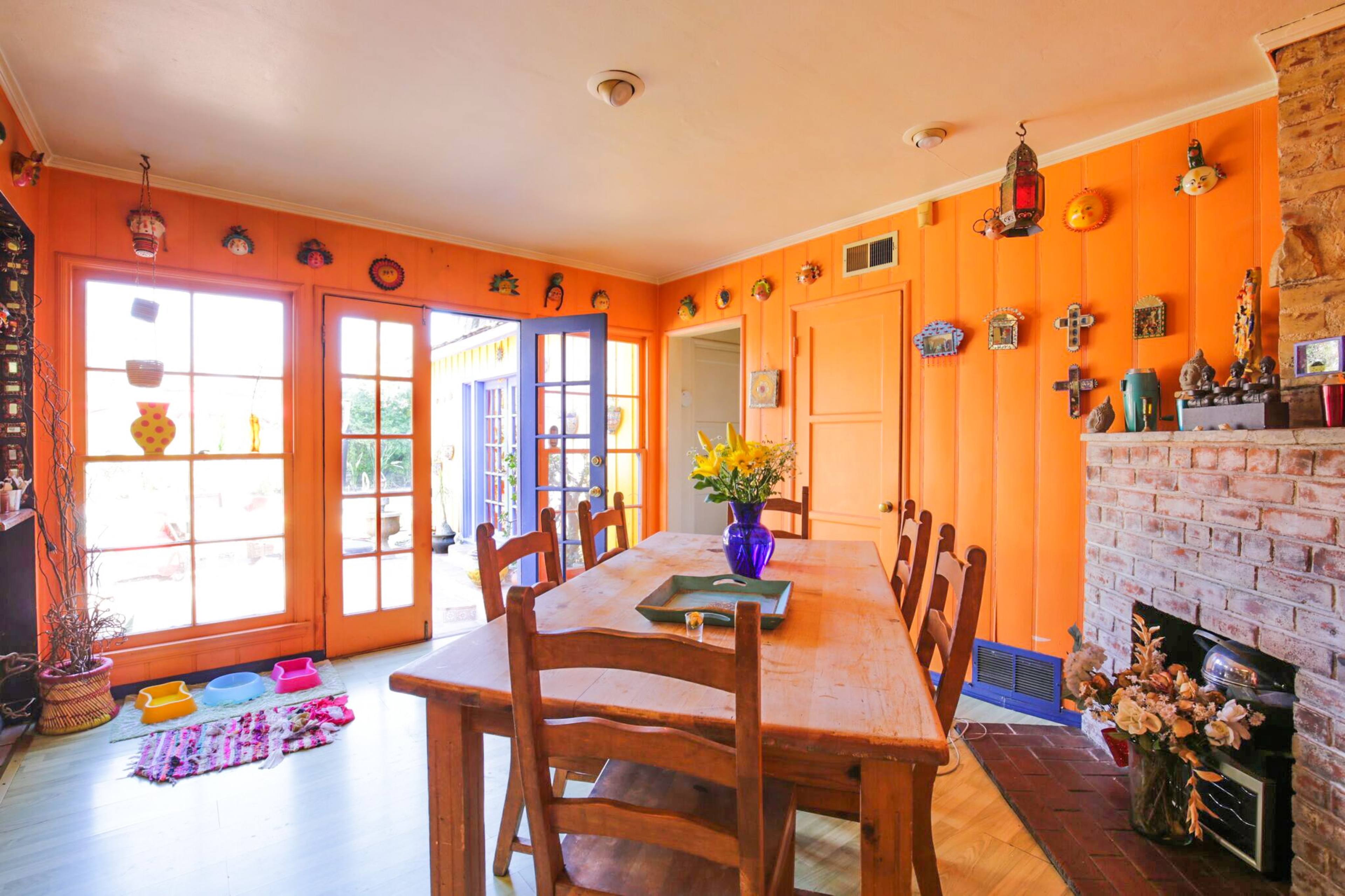 The image shows a brightly colored dining area with orange walls, a wooden table surrounded by chairs, and various decorative items on the walls and shelves.