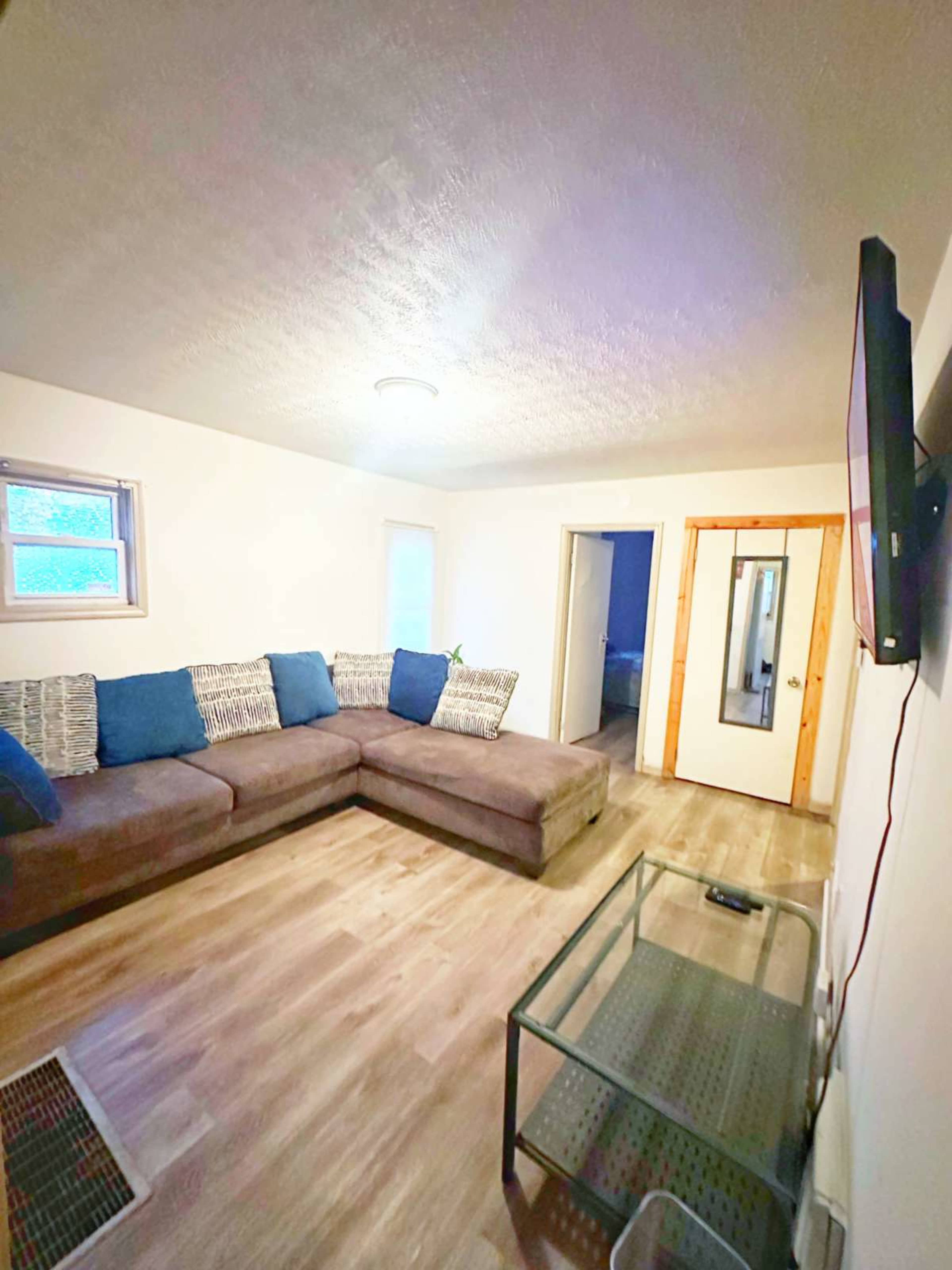 A living room features a sectional sofa with blue throw pillows, a glass coffee table, and a doorway leading to another room.