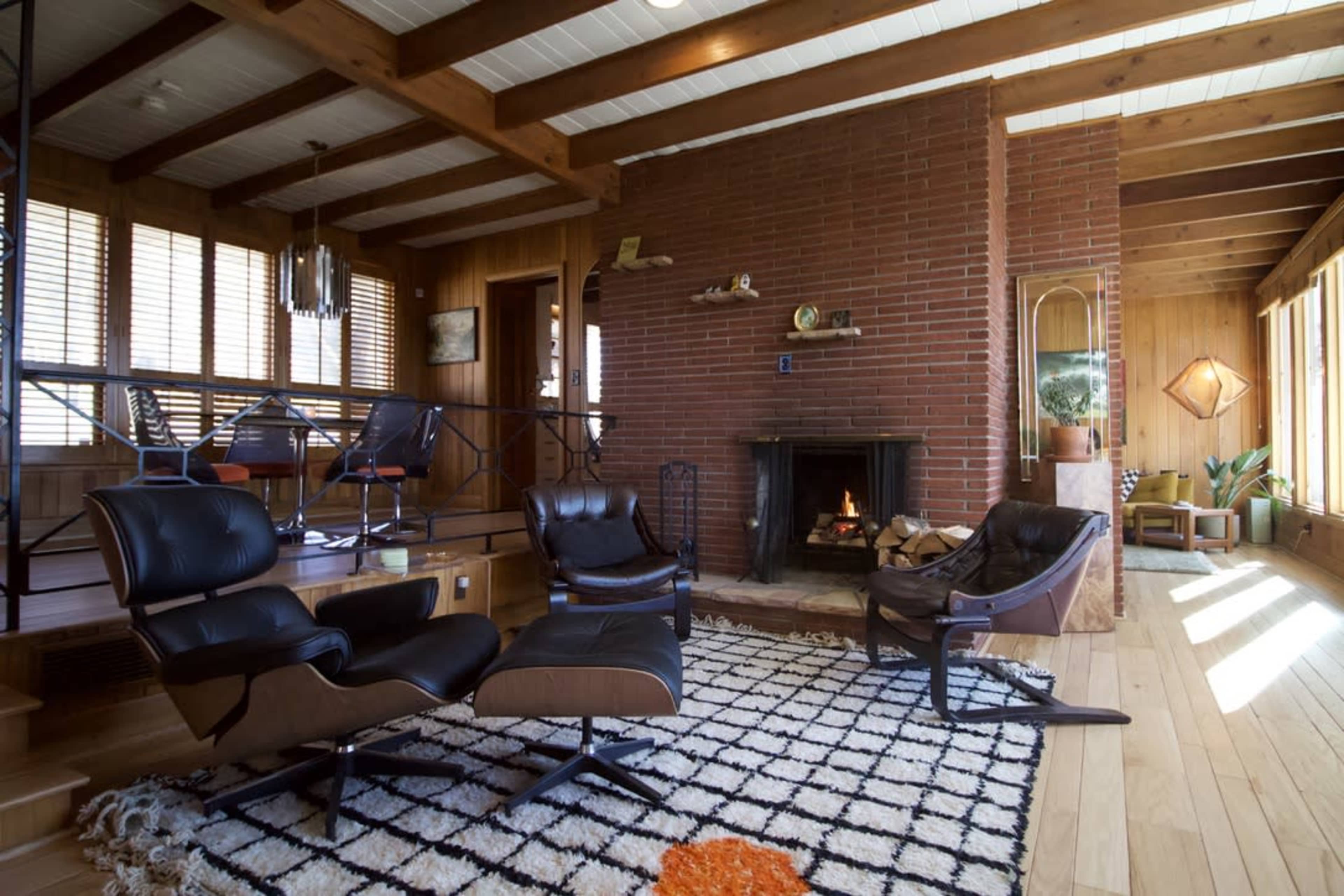 The image shows a wooden-floored living room featuring a brick fireplace, modern chairs, and large windows with wooden beams overhead.