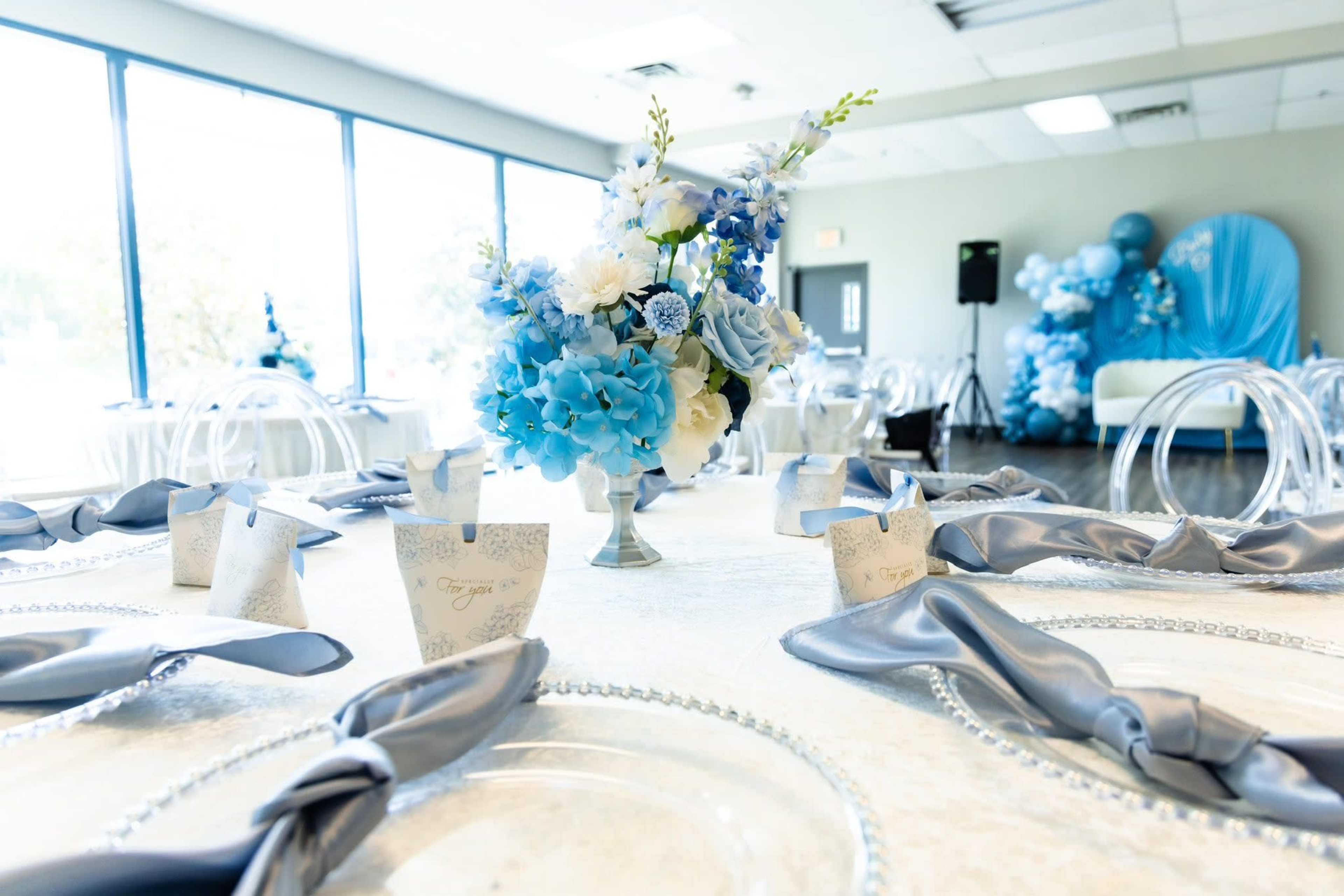 A banquet table is set with clear plates and blue floral centerpieces, surrounded by neatly arranged napkins and decorative cups.