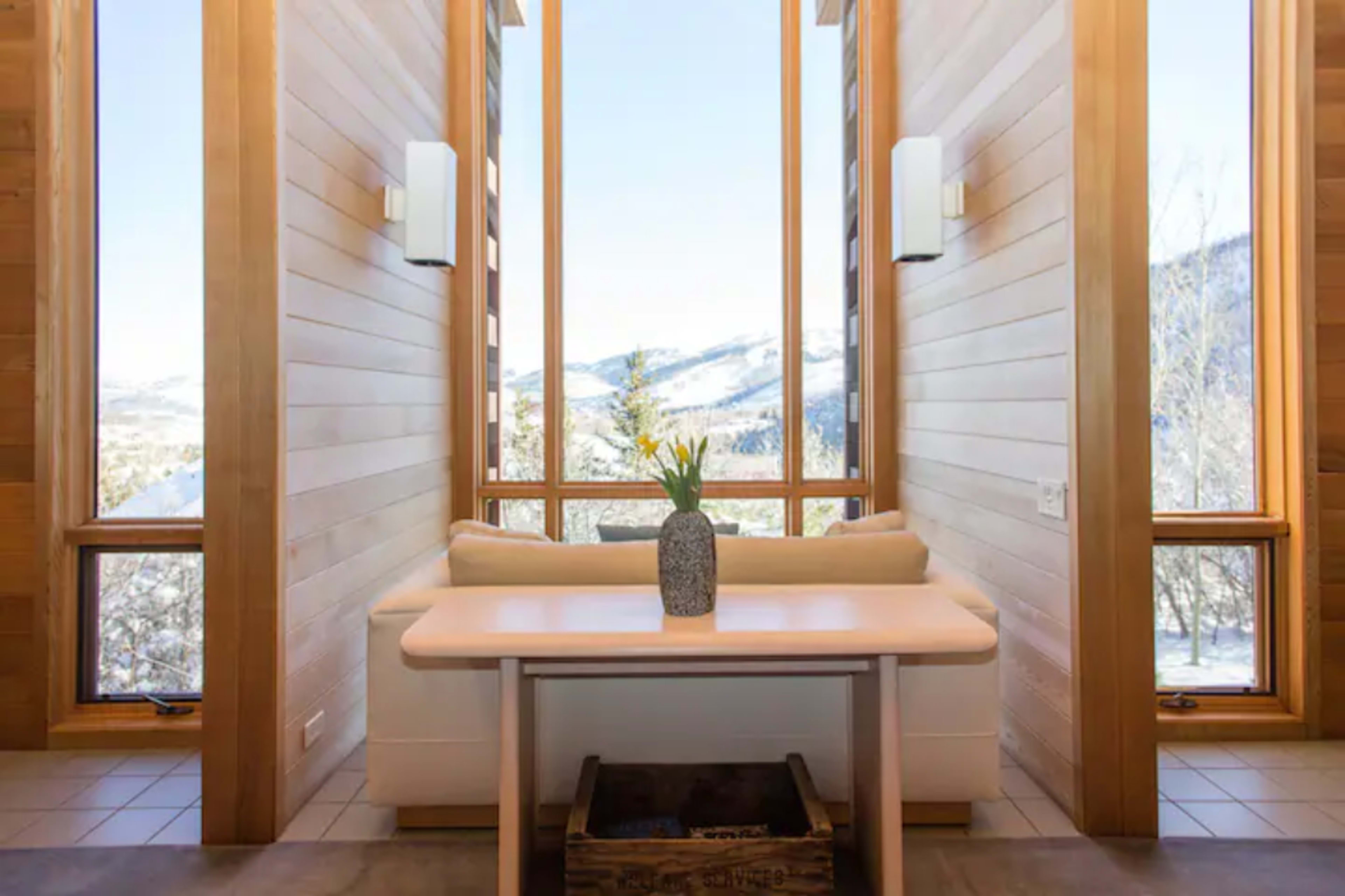 A minimalist room features a table with a vase of flowers, surrounded by large windows that offer a view of snow-capped mountains.