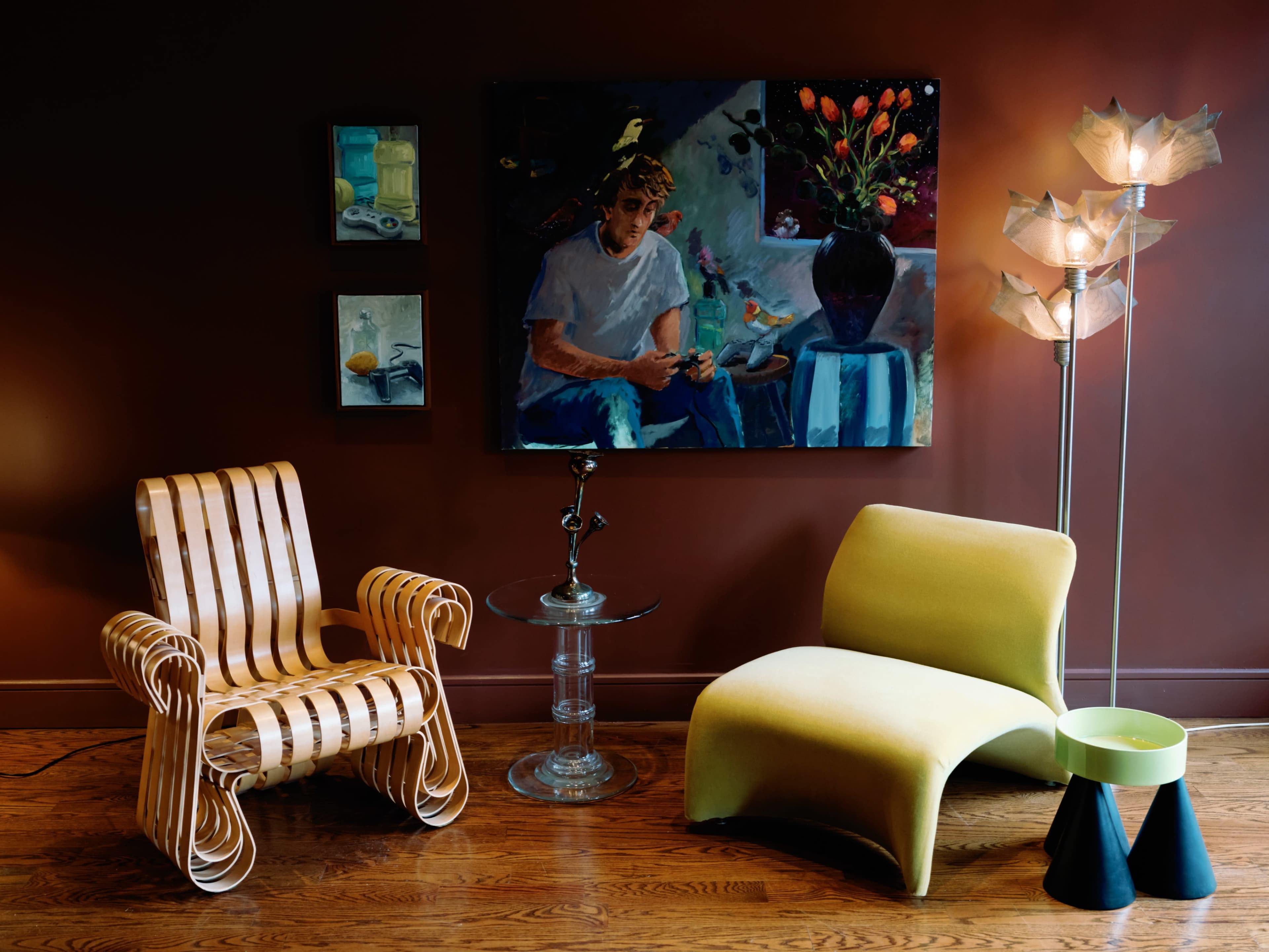 A modern living room features a wooden chair, a curved yellow lounge chair, a standing lamp, and two paintings on a dark brown wall.
