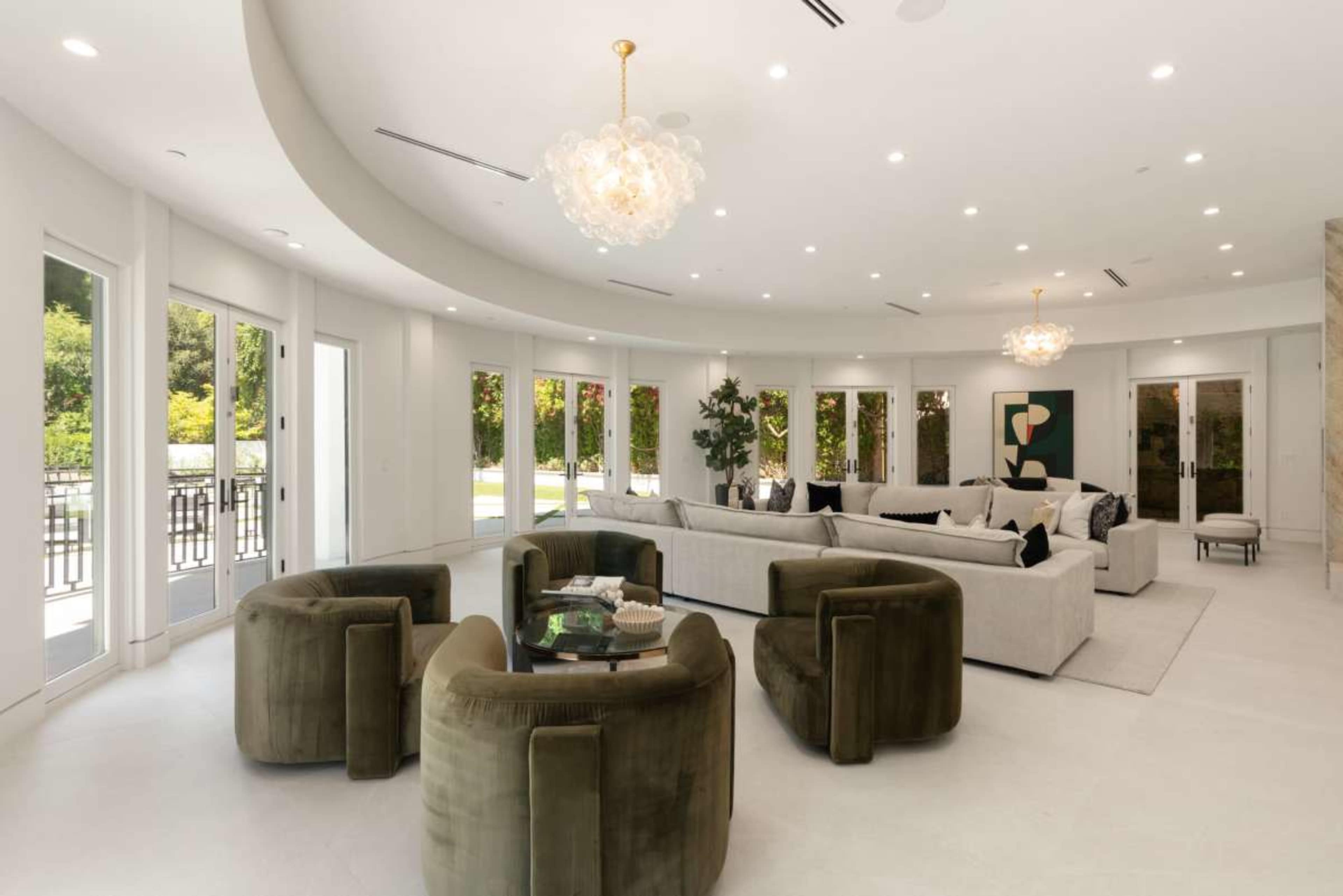 The image shows a spacious modern living room featuring a curved design with a large sectional sofa, plush circular chairs, and multiple glass doors leading to an outdoor area.