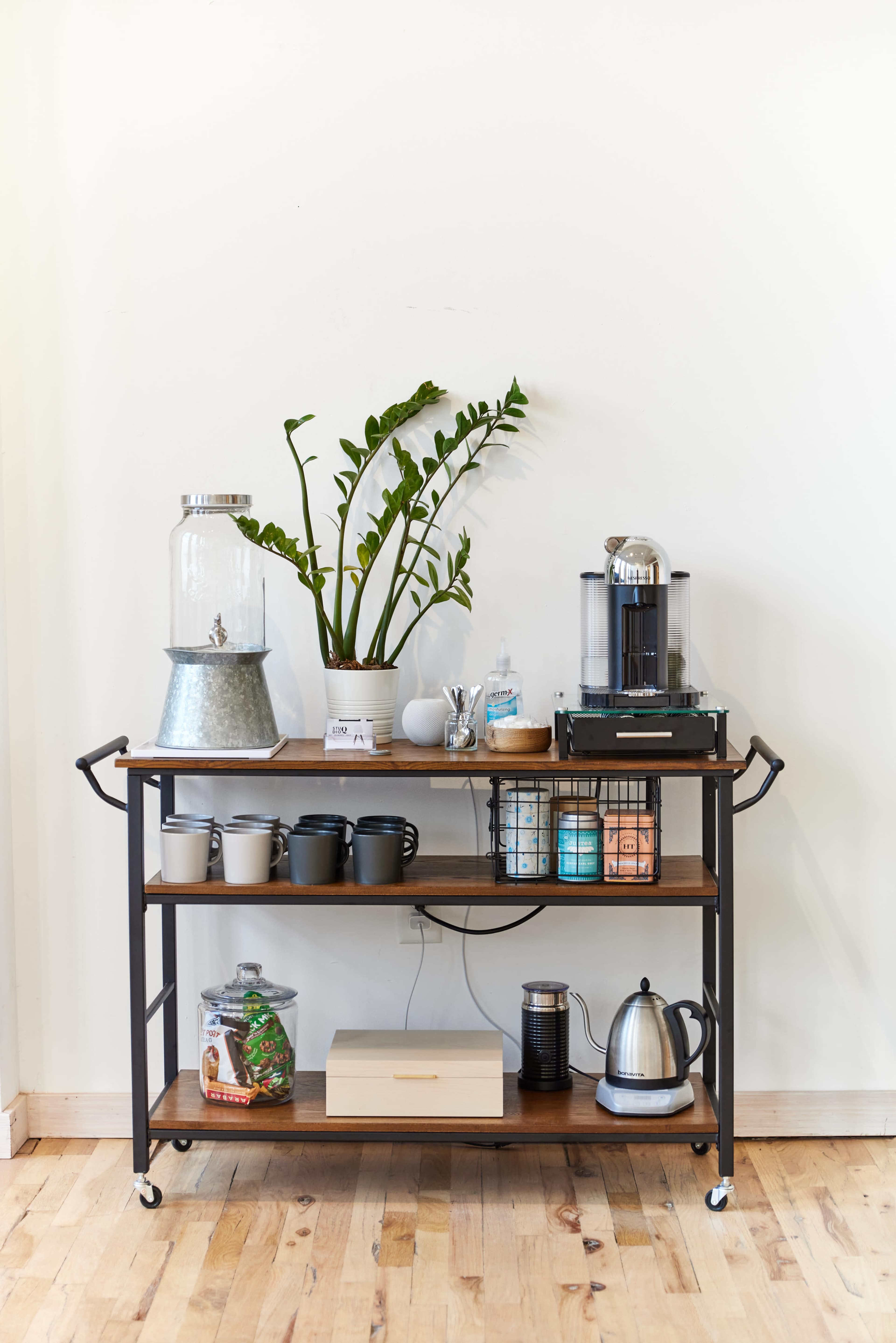 A wooden cart with metal accents holds a coffee maker, water pitcher, mugs, and various containers, positioned against a plain wall with a plant on top.