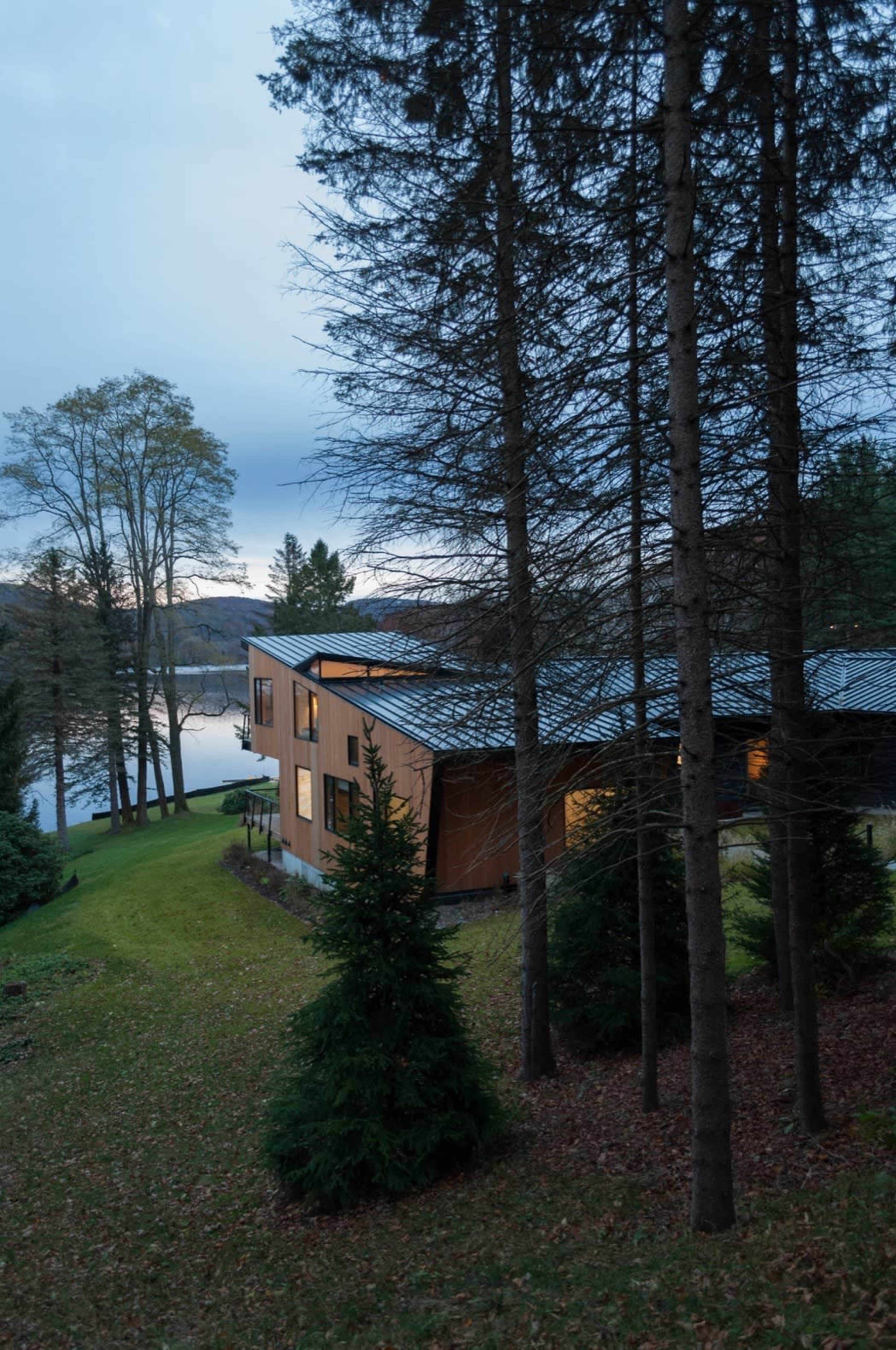 A modern house with large windows is situated among trees overlooking a lake at dusk.