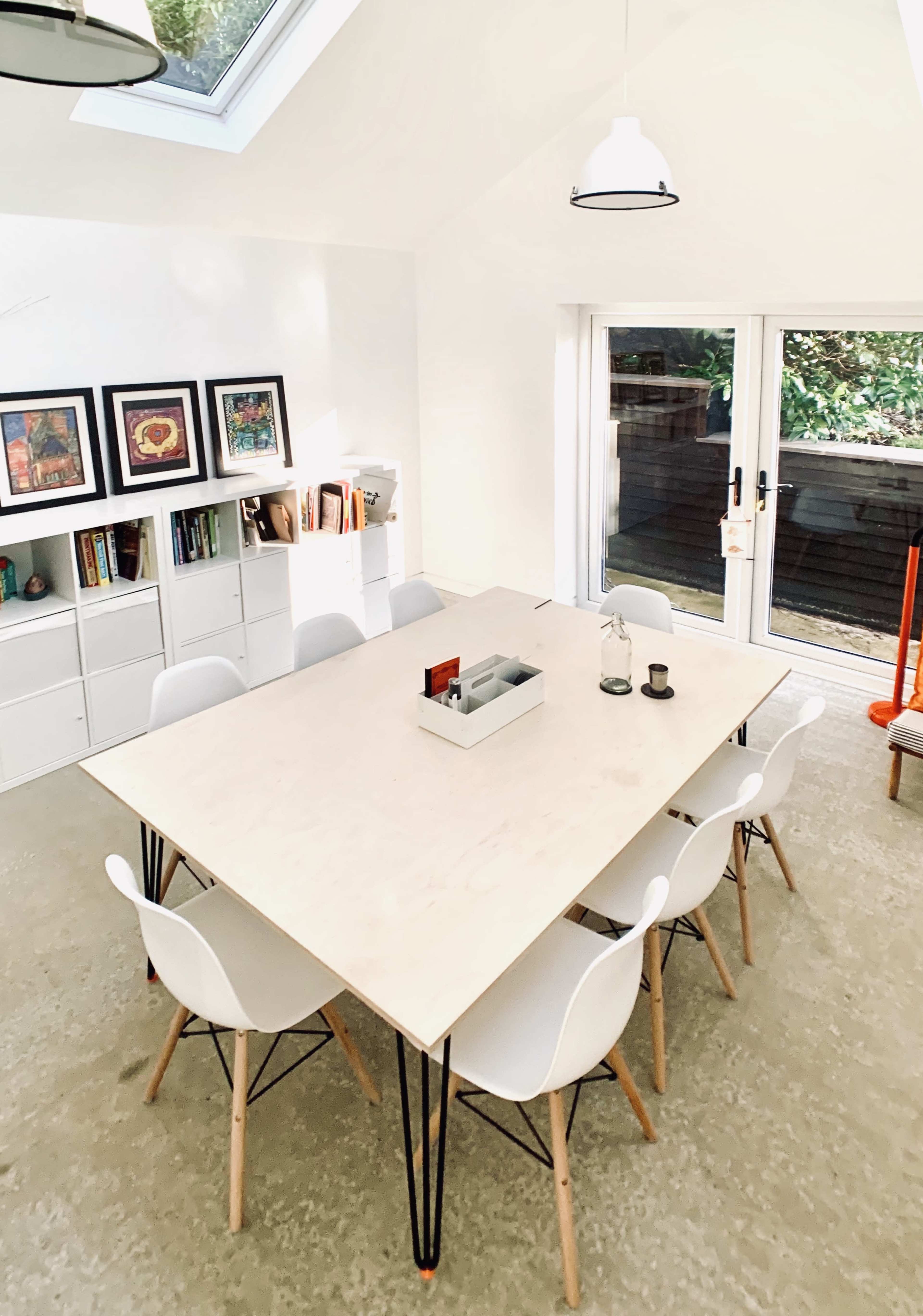 A spacious dining area features a large wooden table surrounded by white chairs, with a bookshelf and large windows letting in natural light.