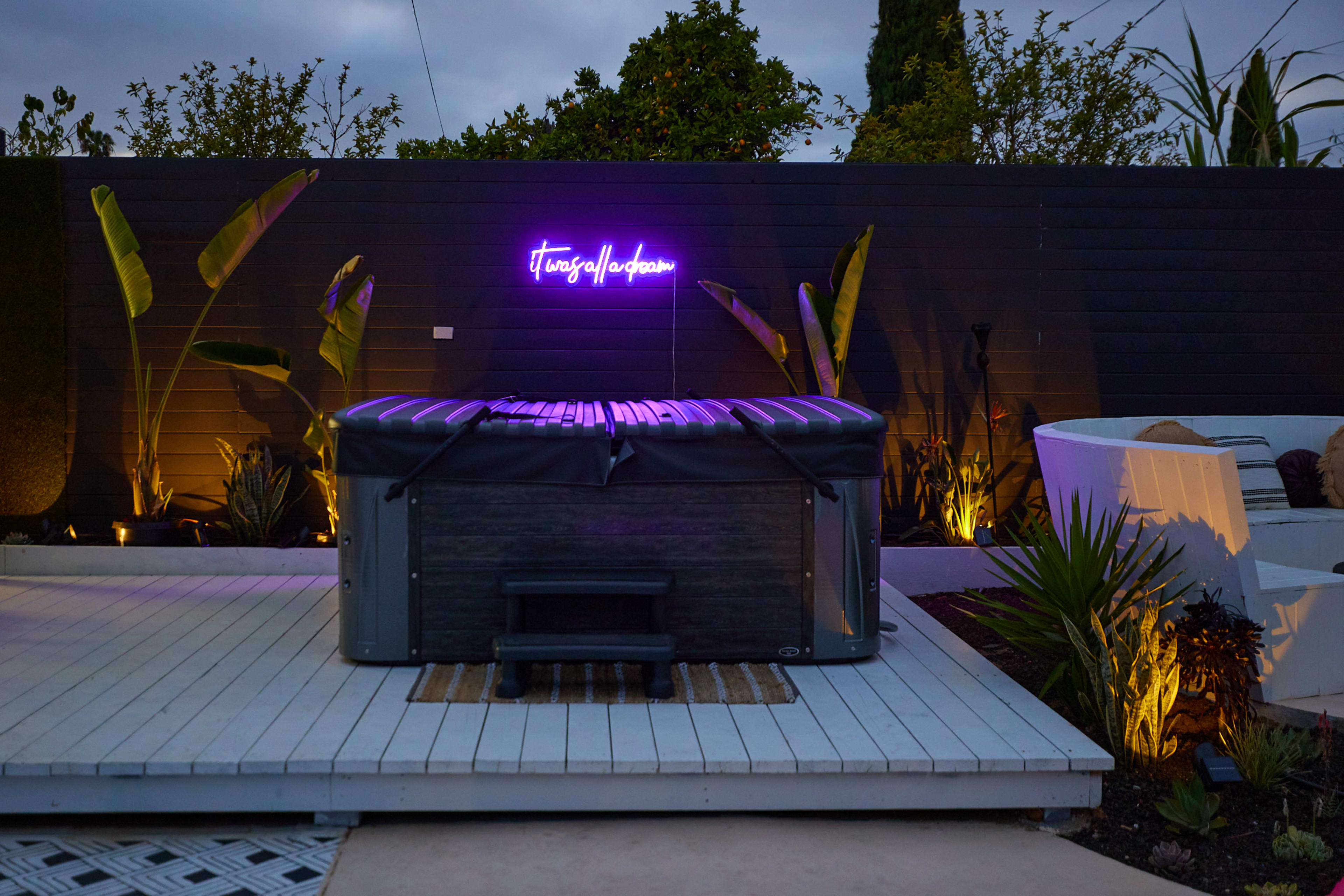 A hot tub with a neon sign sits on a deck surrounded by tropical plants and stylish landscaping.