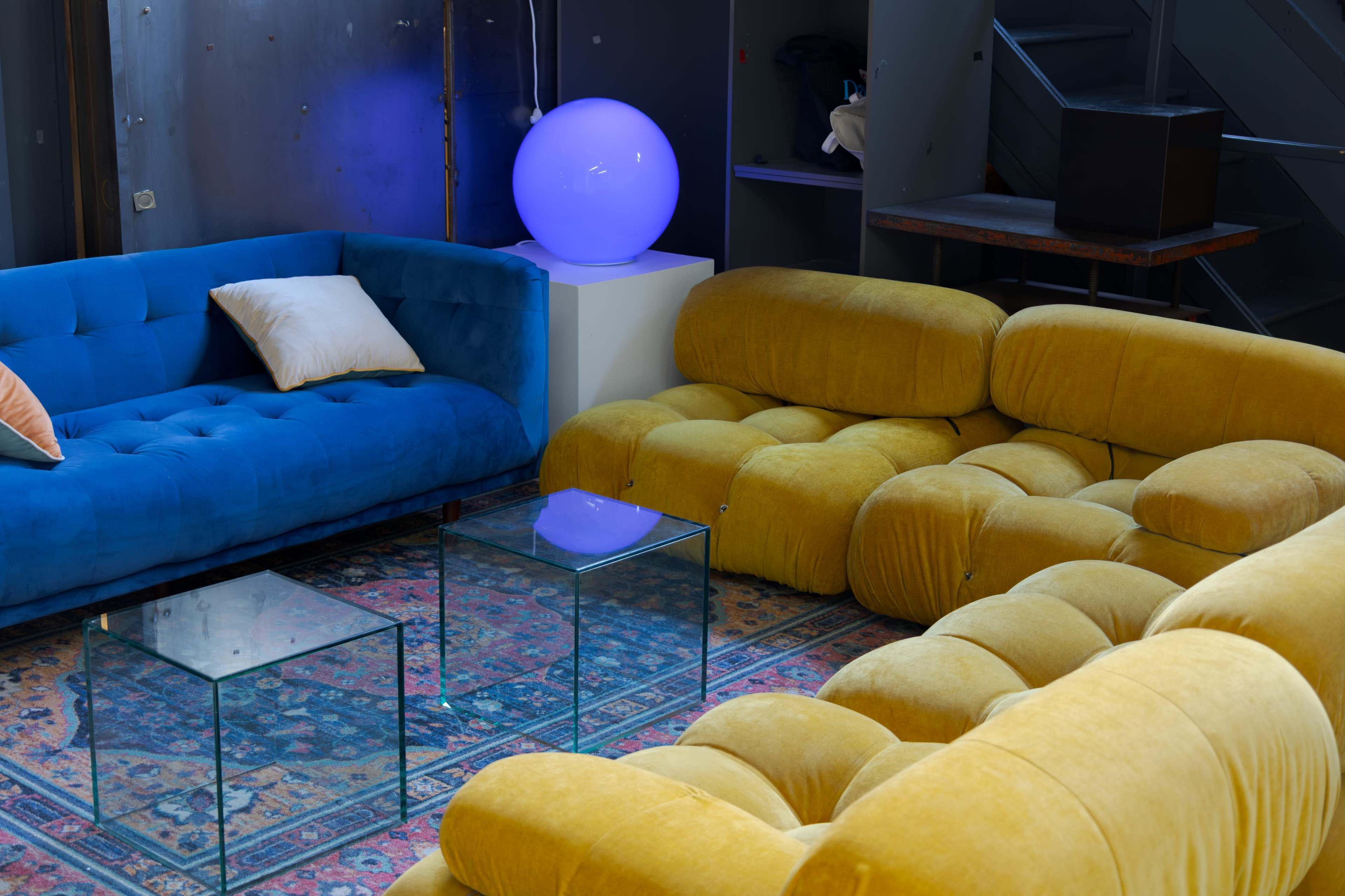 The image shows a modern living space with a blue velvet couch and a yellow plush sectional arranged around two glass tables, accompanied by a glowing spherical lamp on a pedestal.