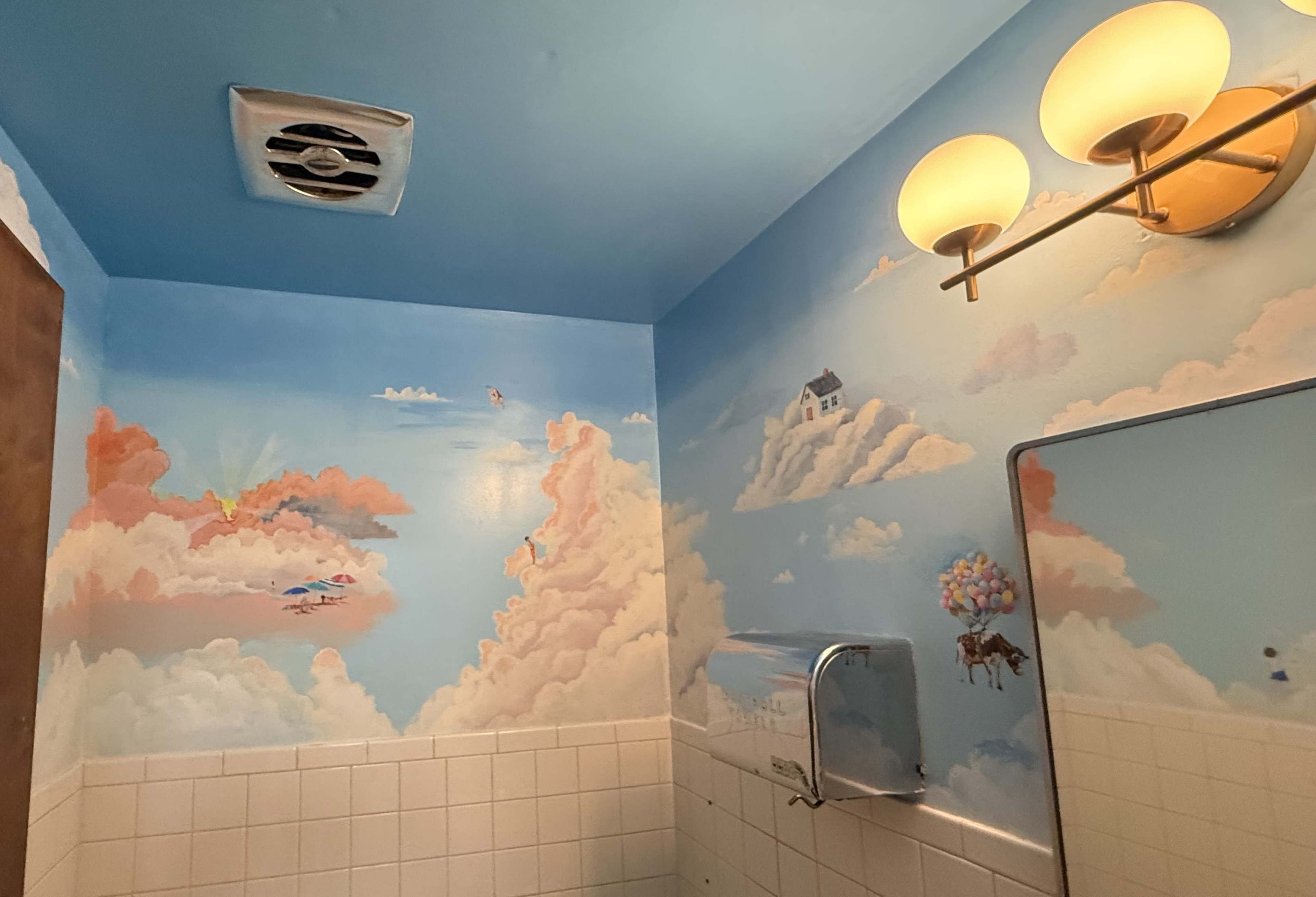 The bathroom features a mural of clouds and a house against a blue sky, complemented by wall-mounted light fixtures and a mirror.