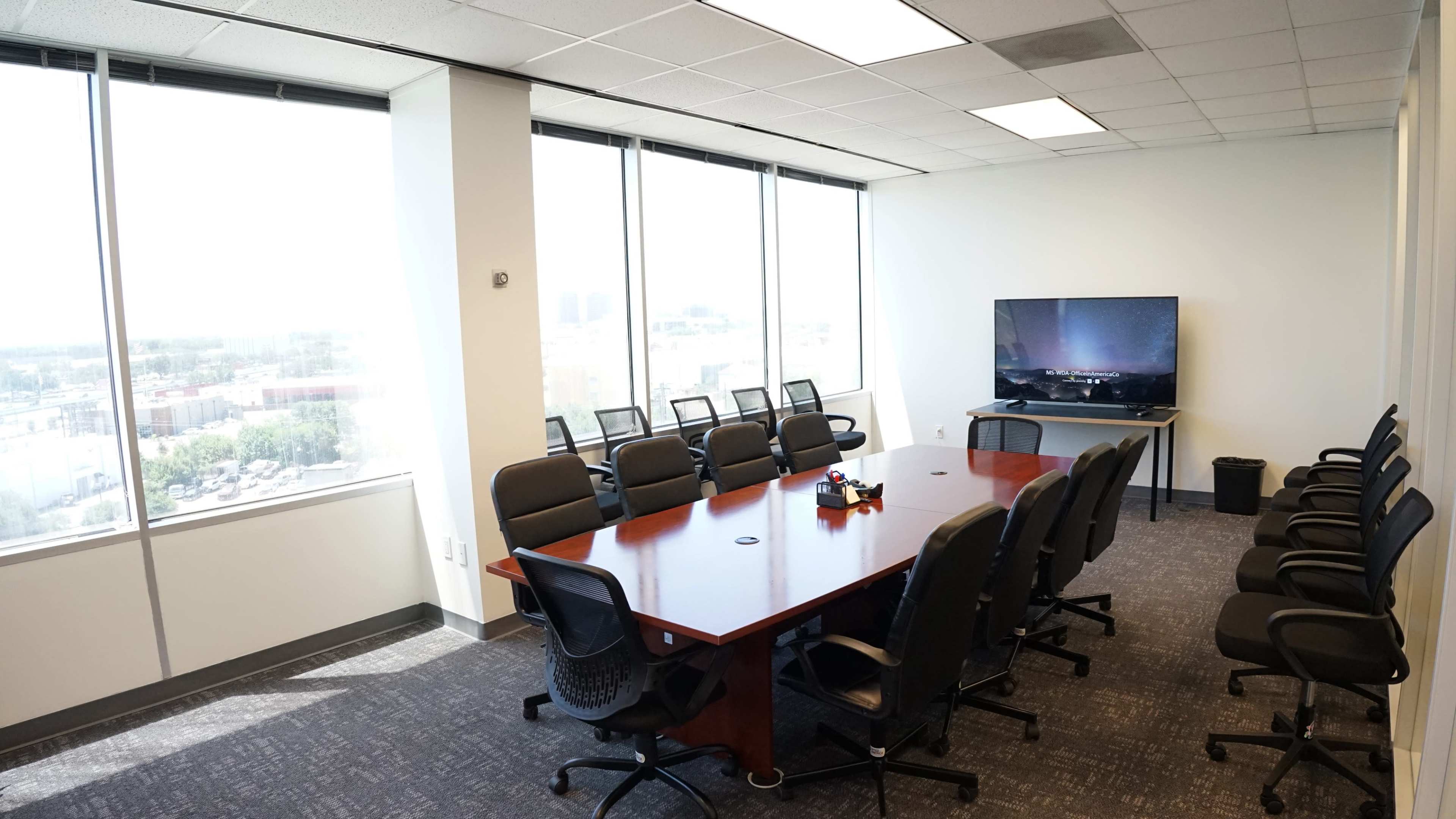 A spacious conference room with a large wooden table and black chairs, overlooking a cityscape through floor-to-ceiling windows.
