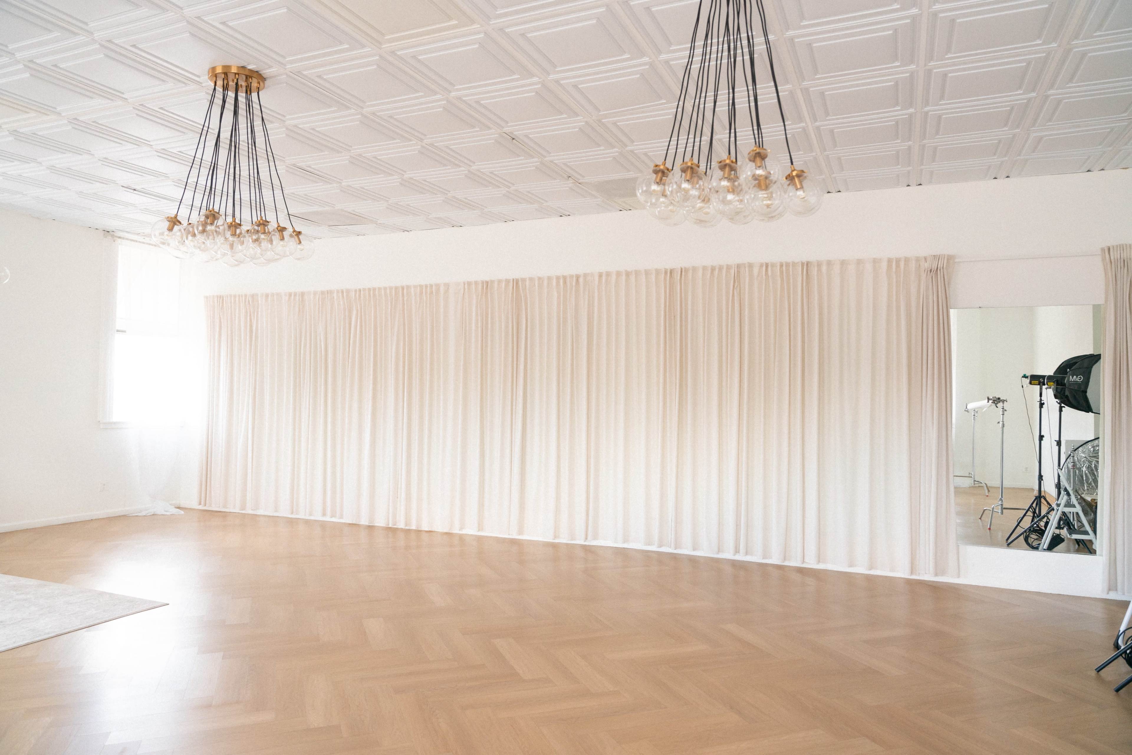 The image shows a spacious room with a light wooden floor, white walls, and elaborate ceiling, featuring multiple pendant lights and sheer curtains along one wall.