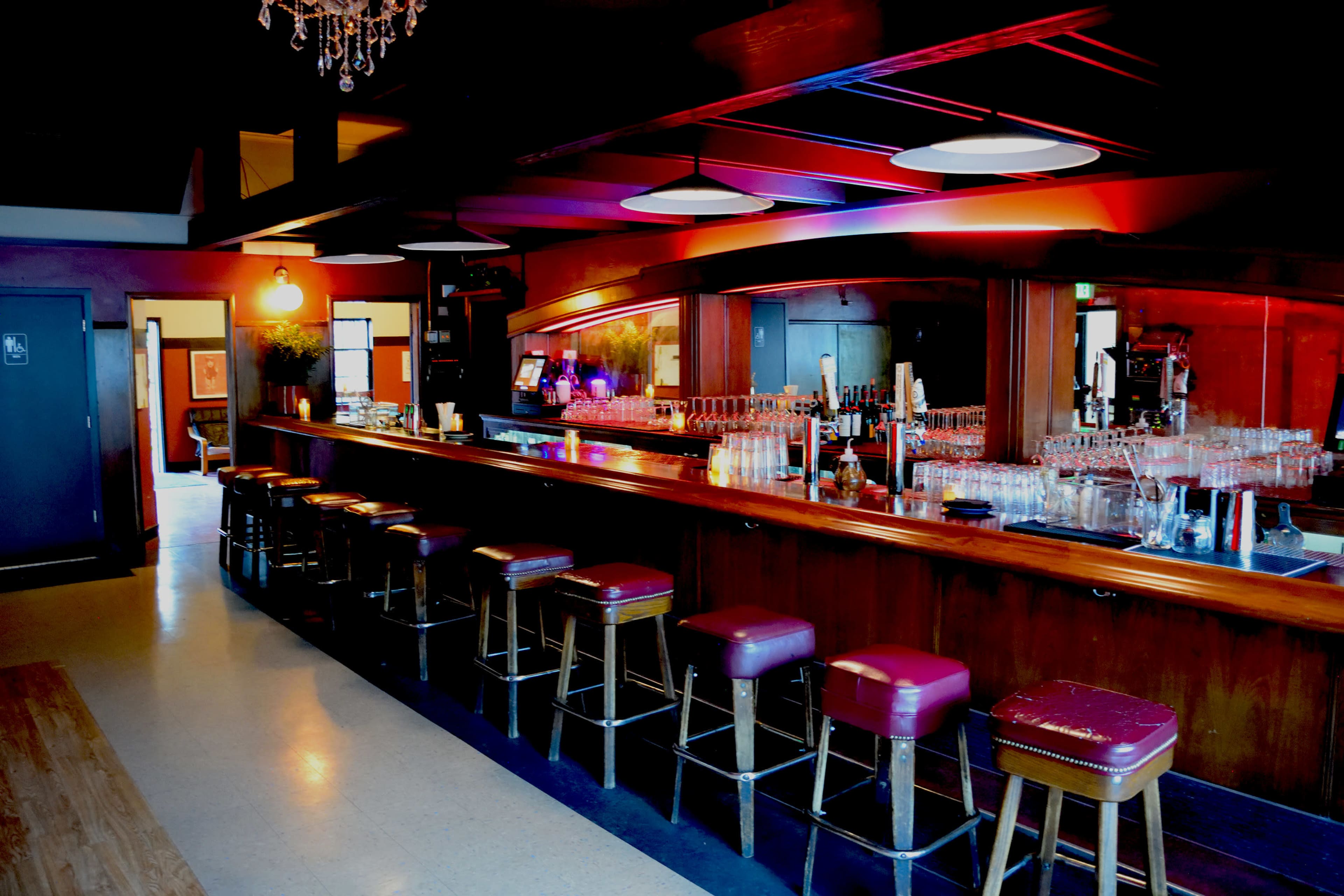 A dimly lit bar with a long wooden counter, several bar stools, and a mirrored back displaying various glassware.