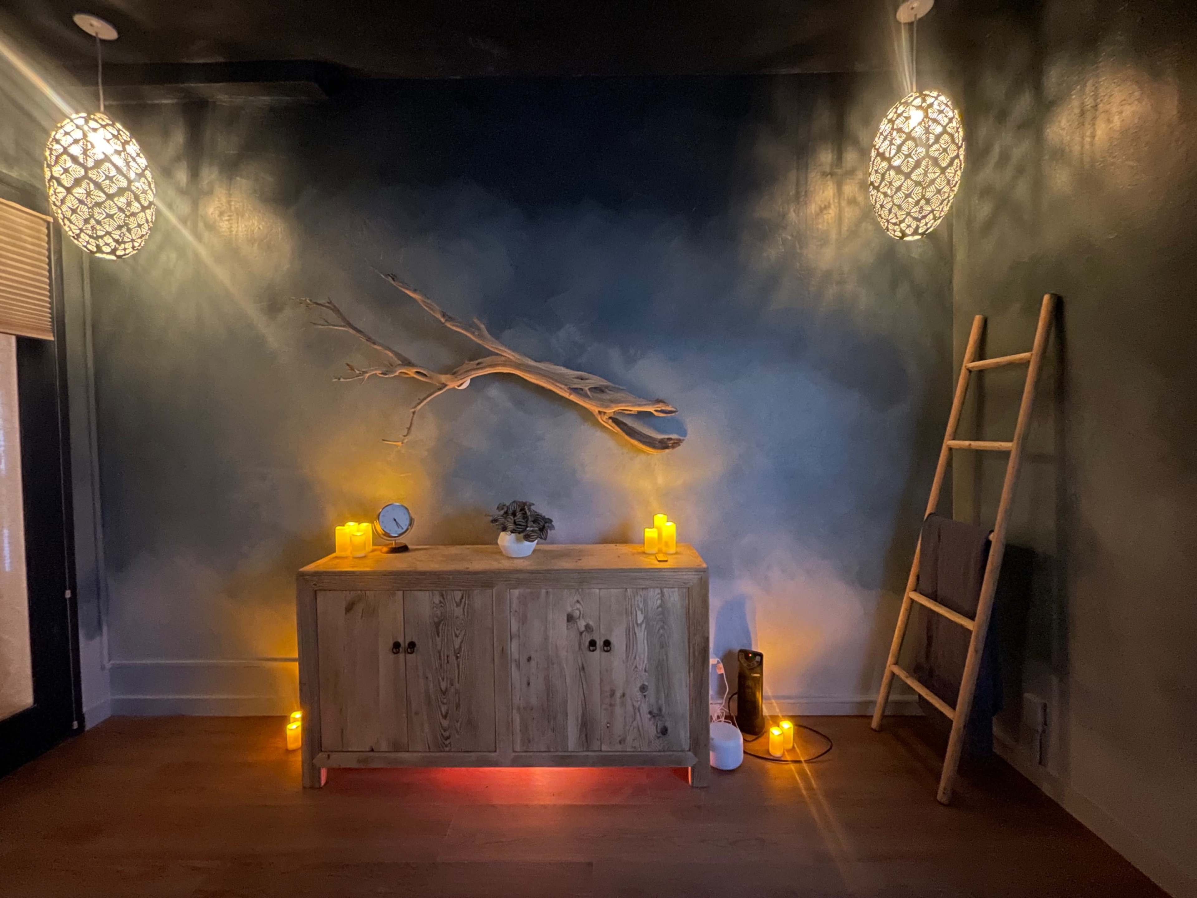 The image shows a softly lit room featuring a wooden sideboard with candles, a small plant, and a decorative wall with a branch, along with a ladder leaning against the wall.