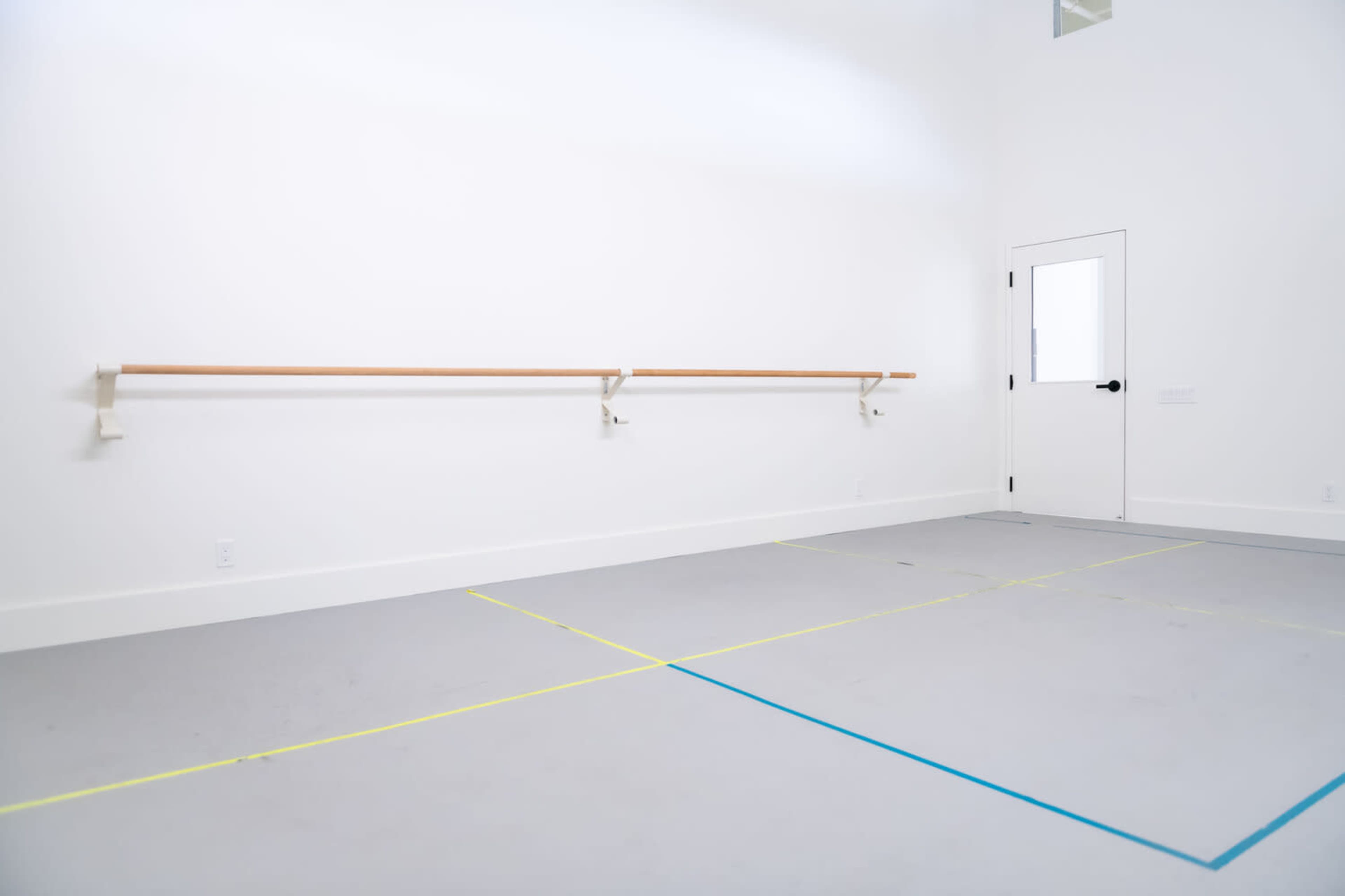 A spacious dance studio featuring a plain white wall with a wooden ballet bar and a door on the side.