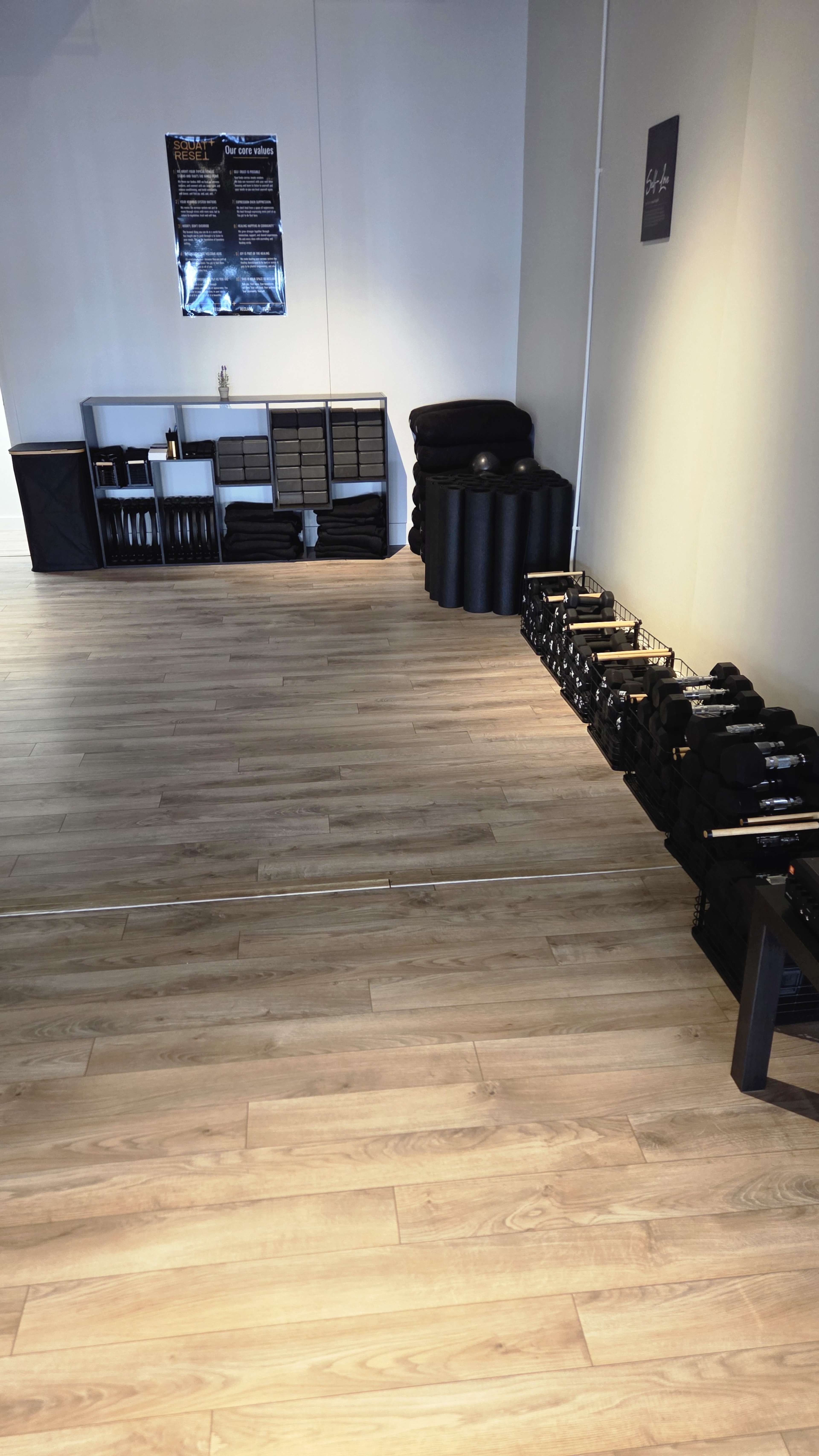 The image shows a spacious, empty room with wooden floors, storage shelves on one side, and various workout equipment, including weights and foam rollers, neatly arranged along the walls.