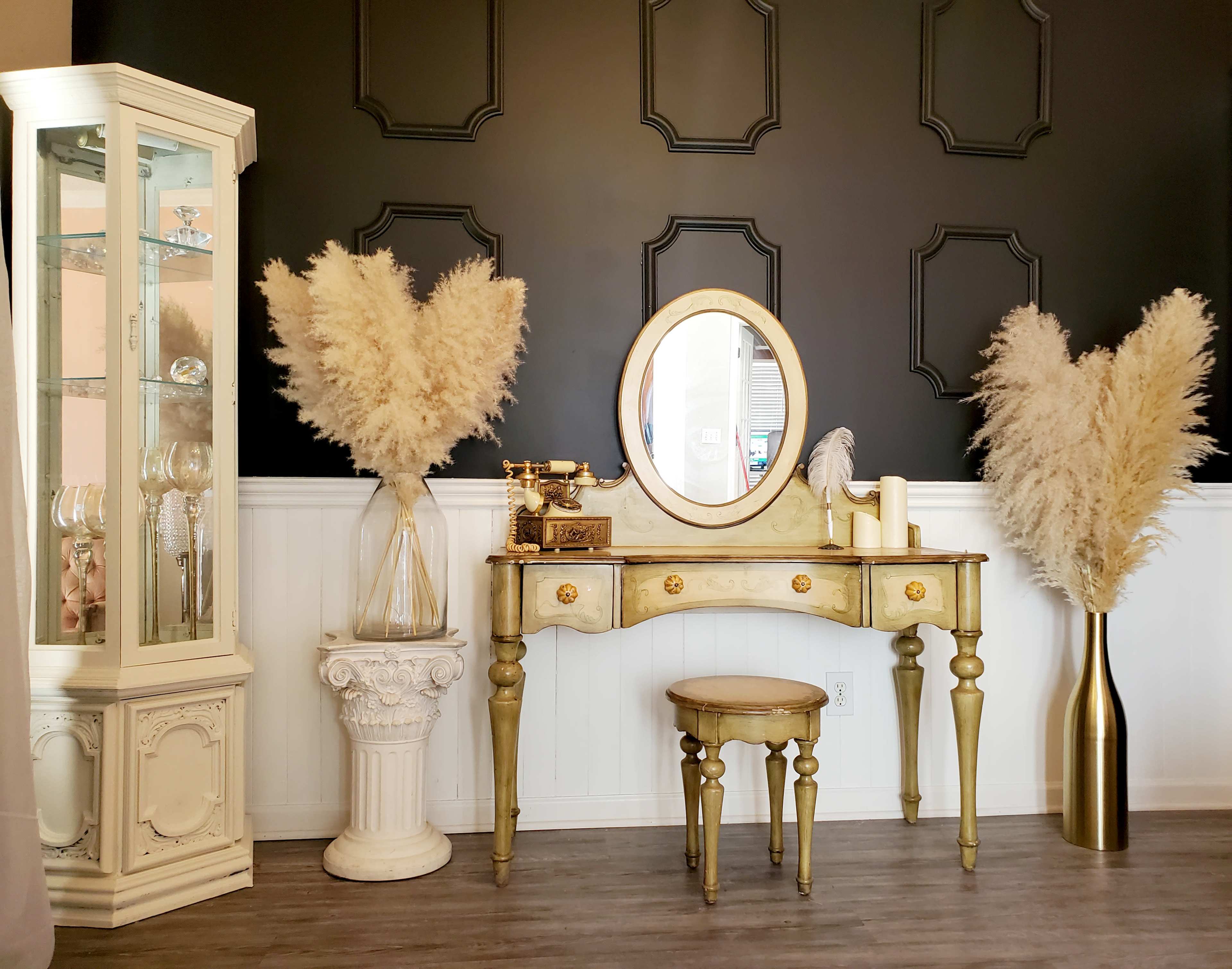 The image features a decorated corner of a room with a vintage vanity table, a round mirror, and two large vases containing pampas grass, set against a black wall with elegant paneling.