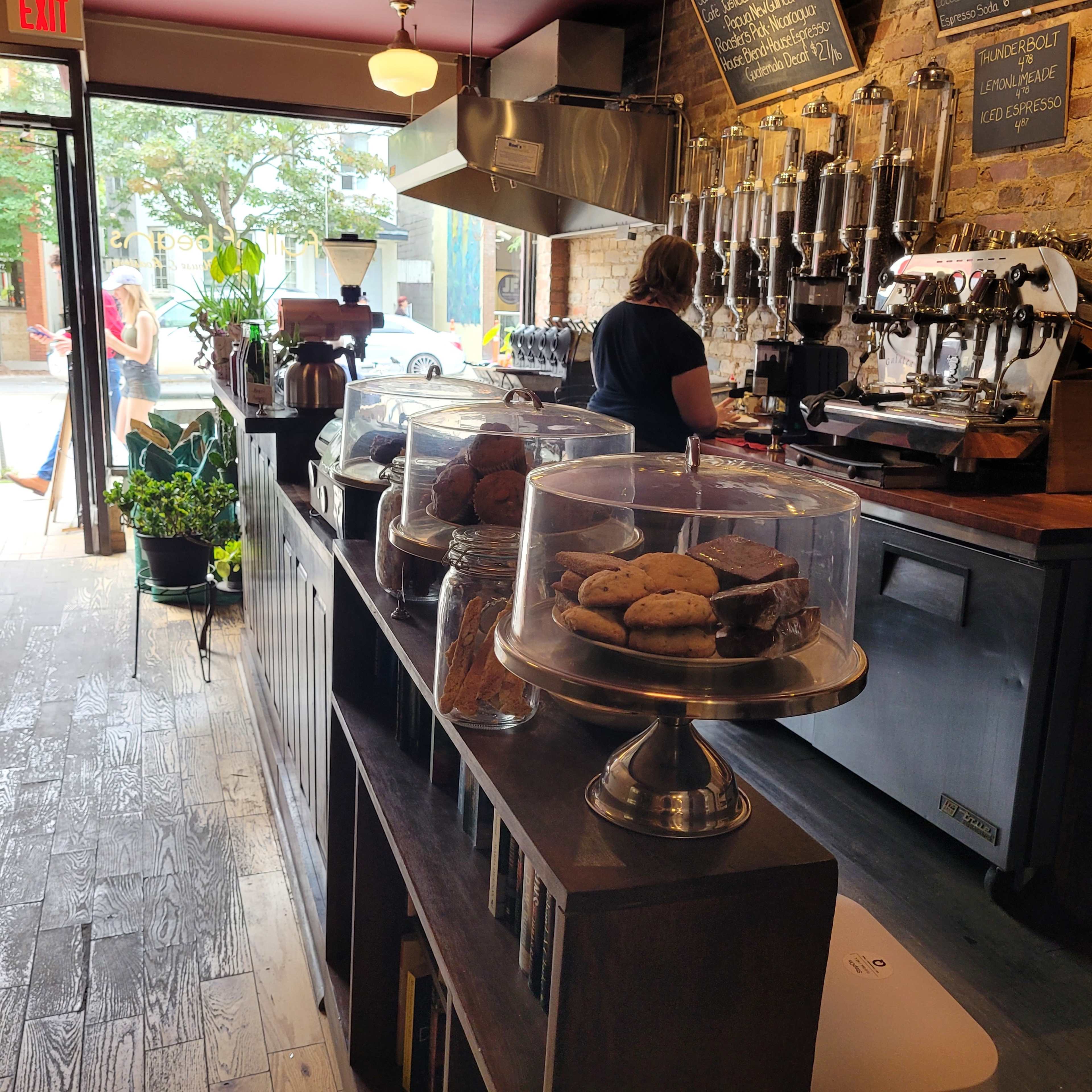 A coffee shop interior features a display of cookies under glass domes, with a barista preparing drinks behind the counter, and a view of the street through the open door.