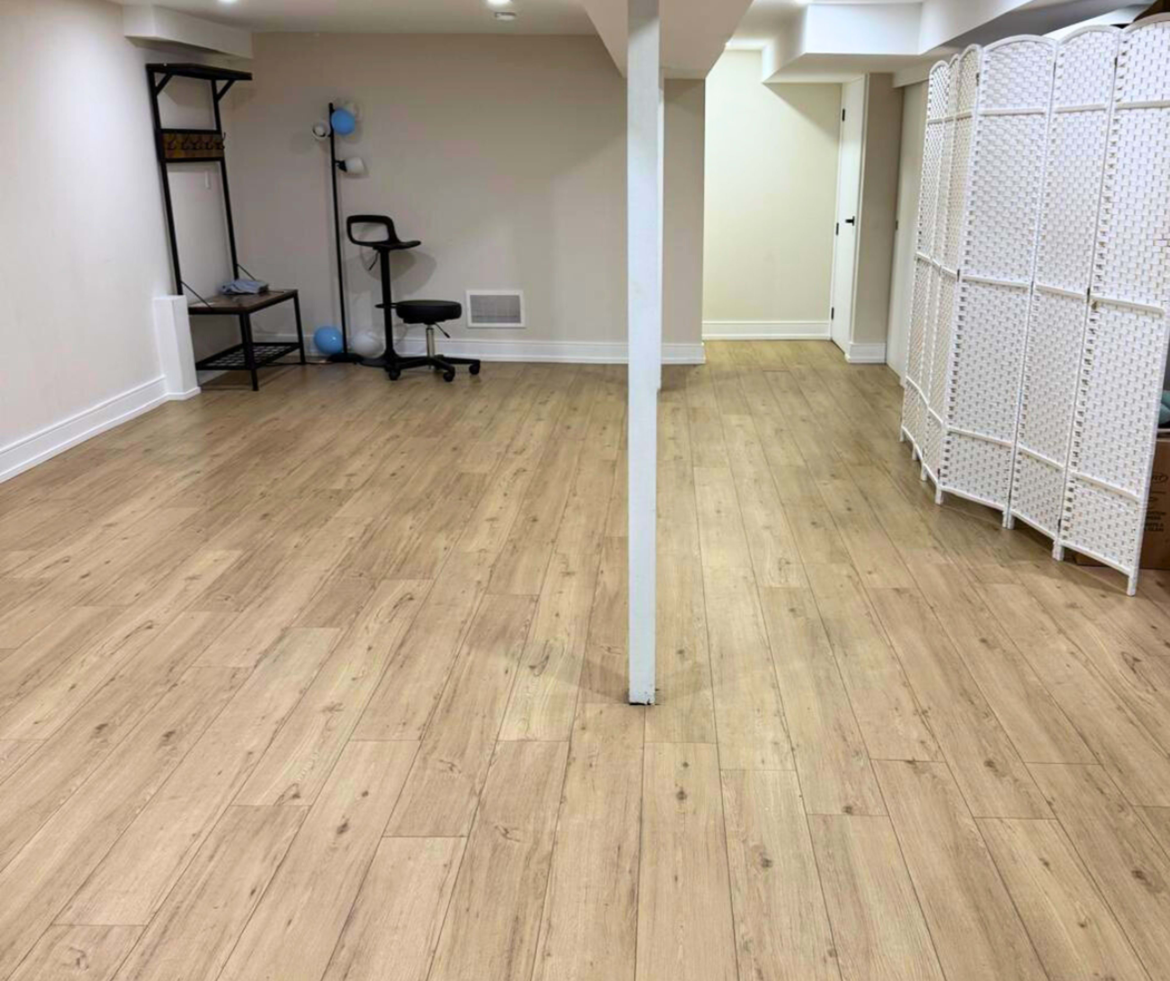 The image shows a spacious room with wooden flooring, featuring a black office chair and a room divider on one side.