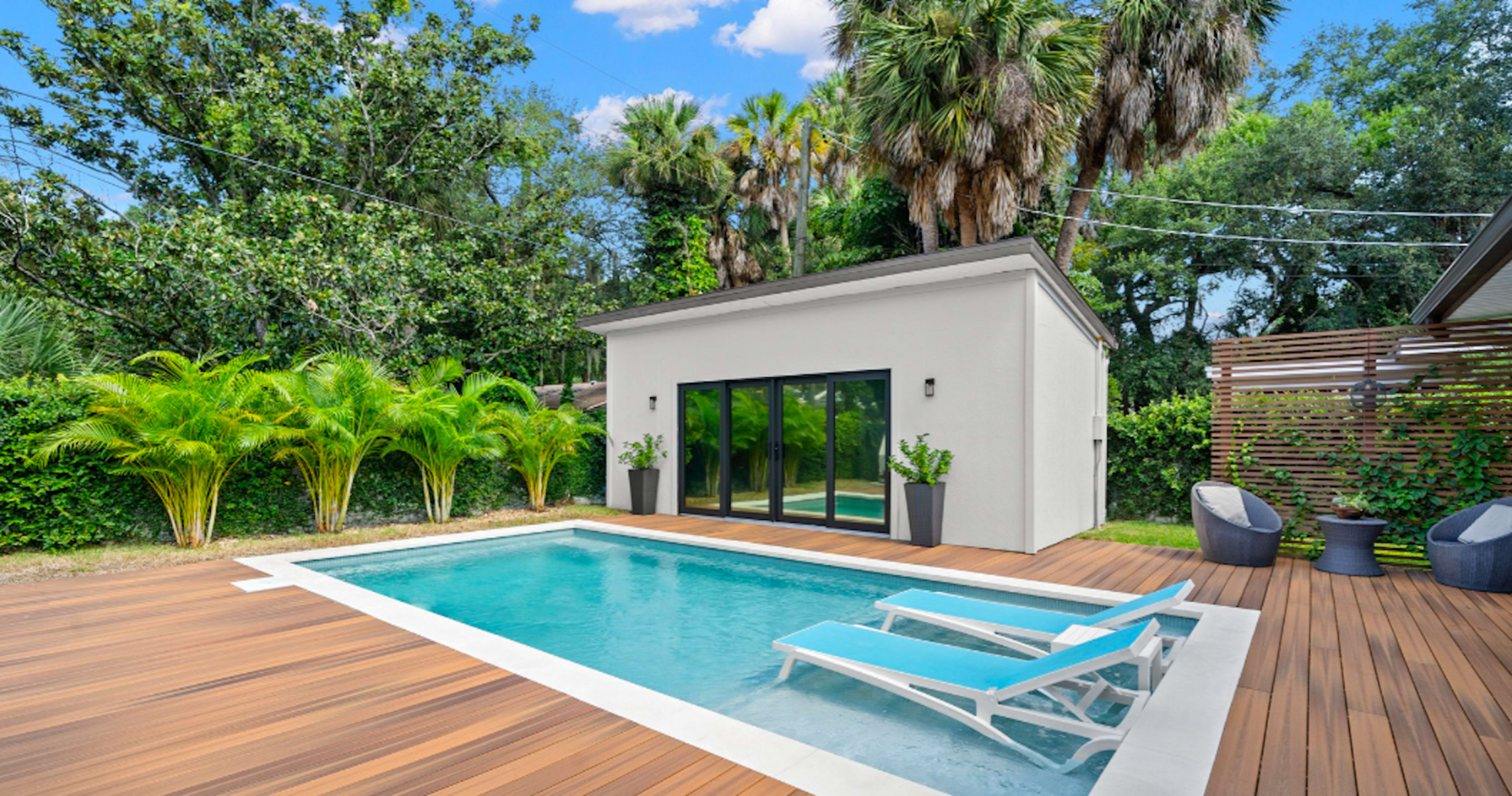 A small pool surrounded by wooden decking, with lounge chairs and a modern building in a lush, tropical garden.