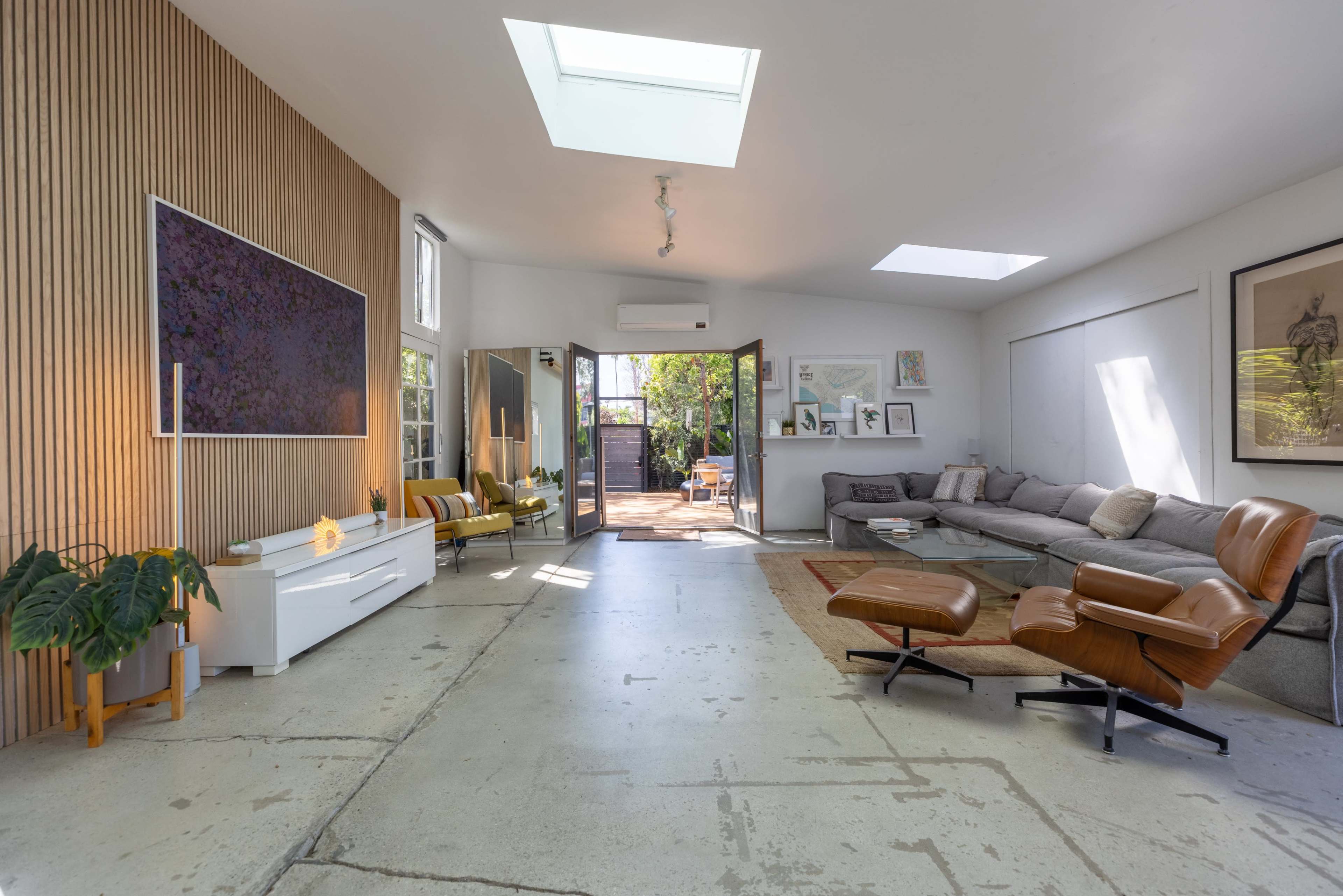 The image shows a modern living room featuring a large TV mounted on a wooden wall, a sectional sofa, and a skylight illuminating the space.