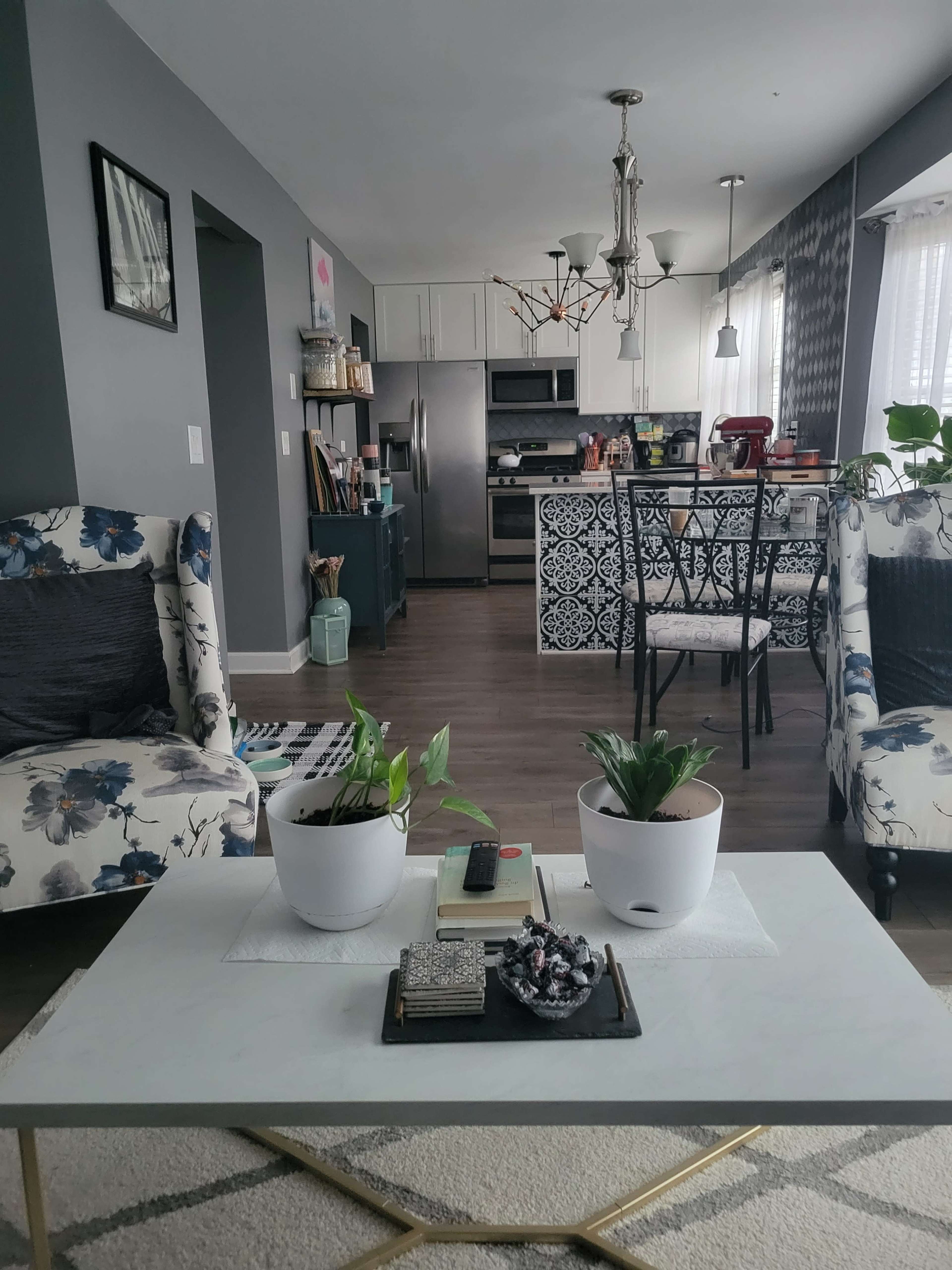 A modern living room with floral-patterned chairs, a coffee table with plants and books, and an open kitchen in the background.