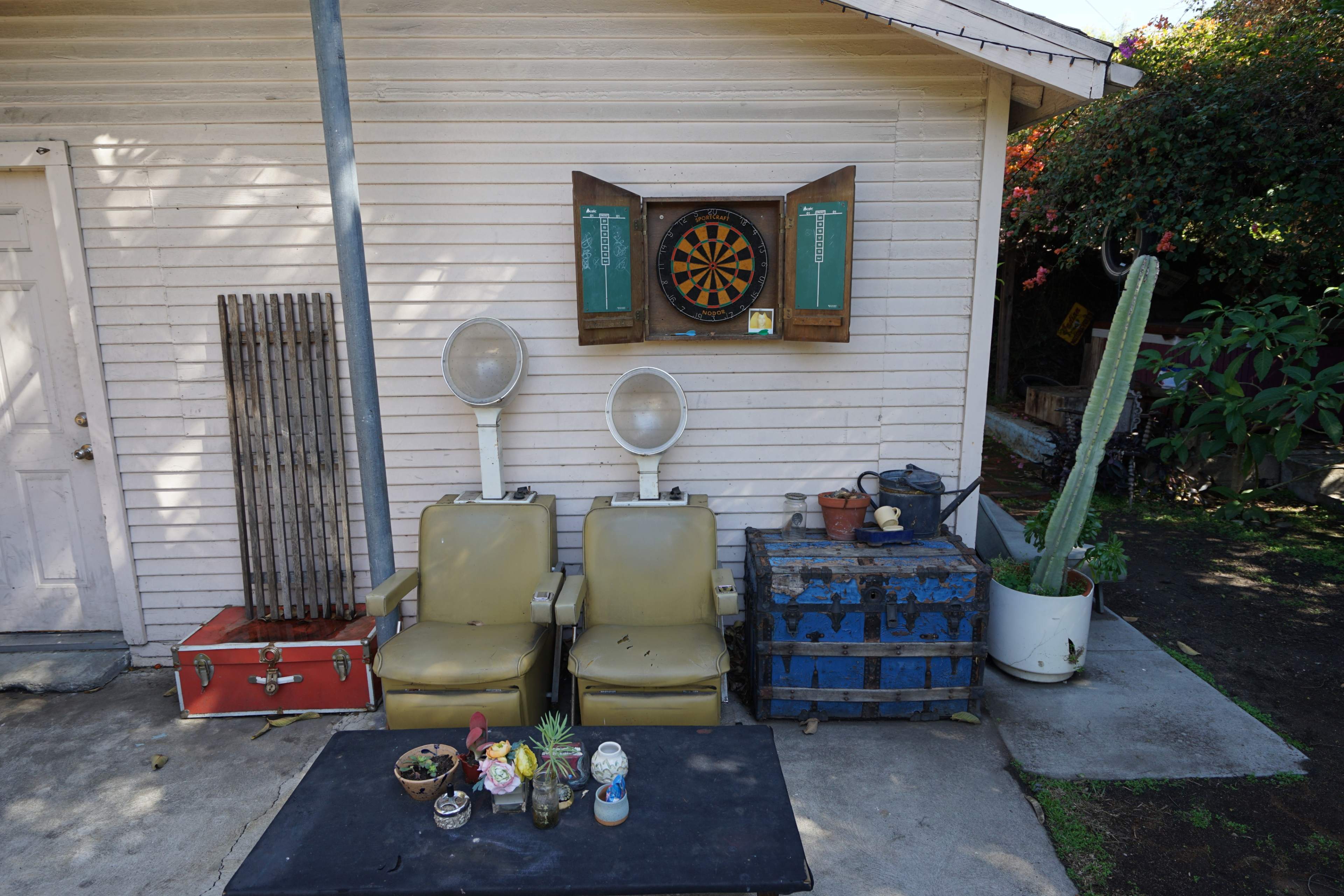 Two vintage chairs are positioned in front of a light-colored shed, with a dartboard mounted above them and a variety of potted plants and objects on a nearby table.