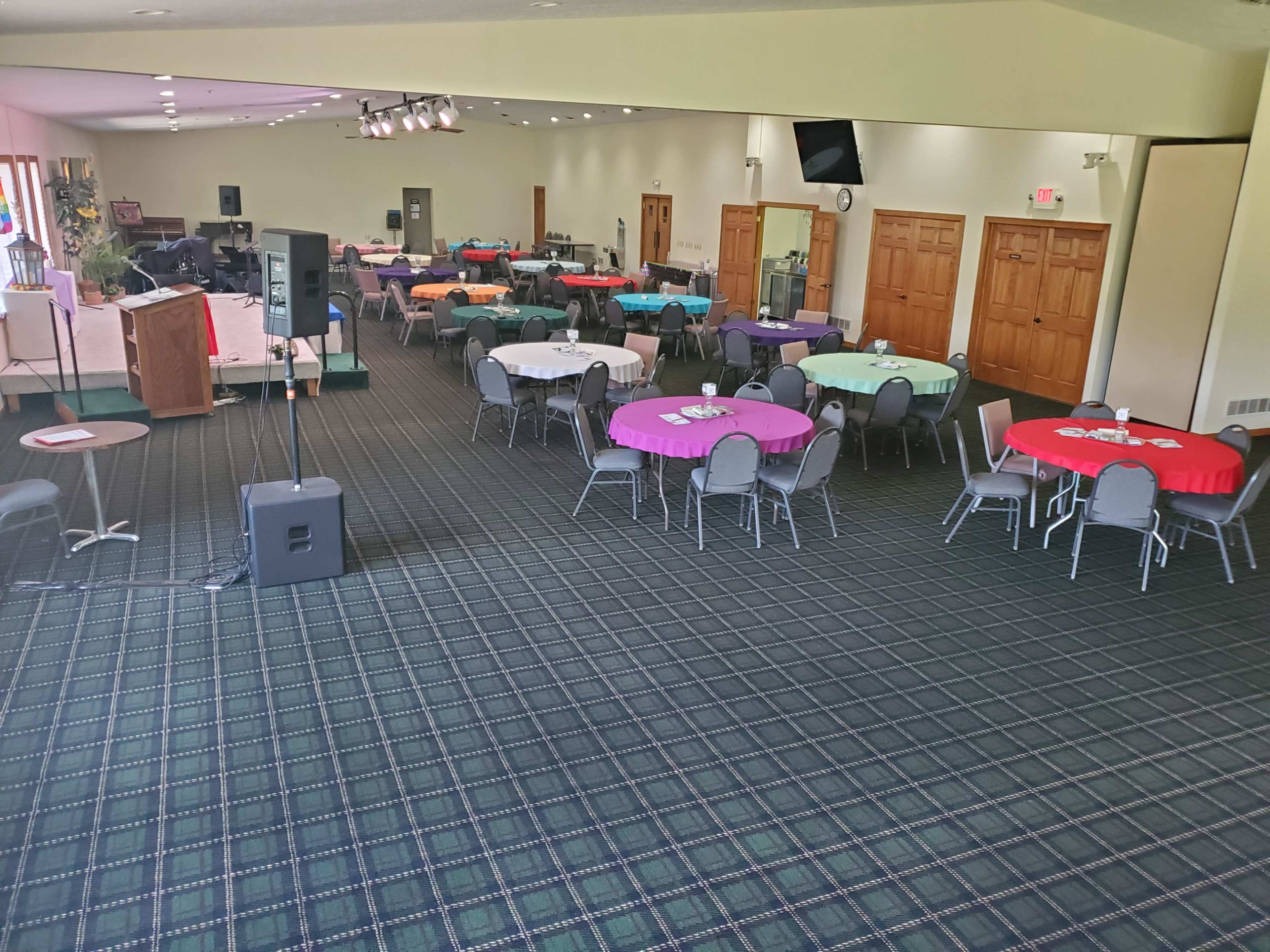 A large room with several round tables covered in colorful tablecloths is set up for an event, with a podium and speaker equipment positioned at the front.