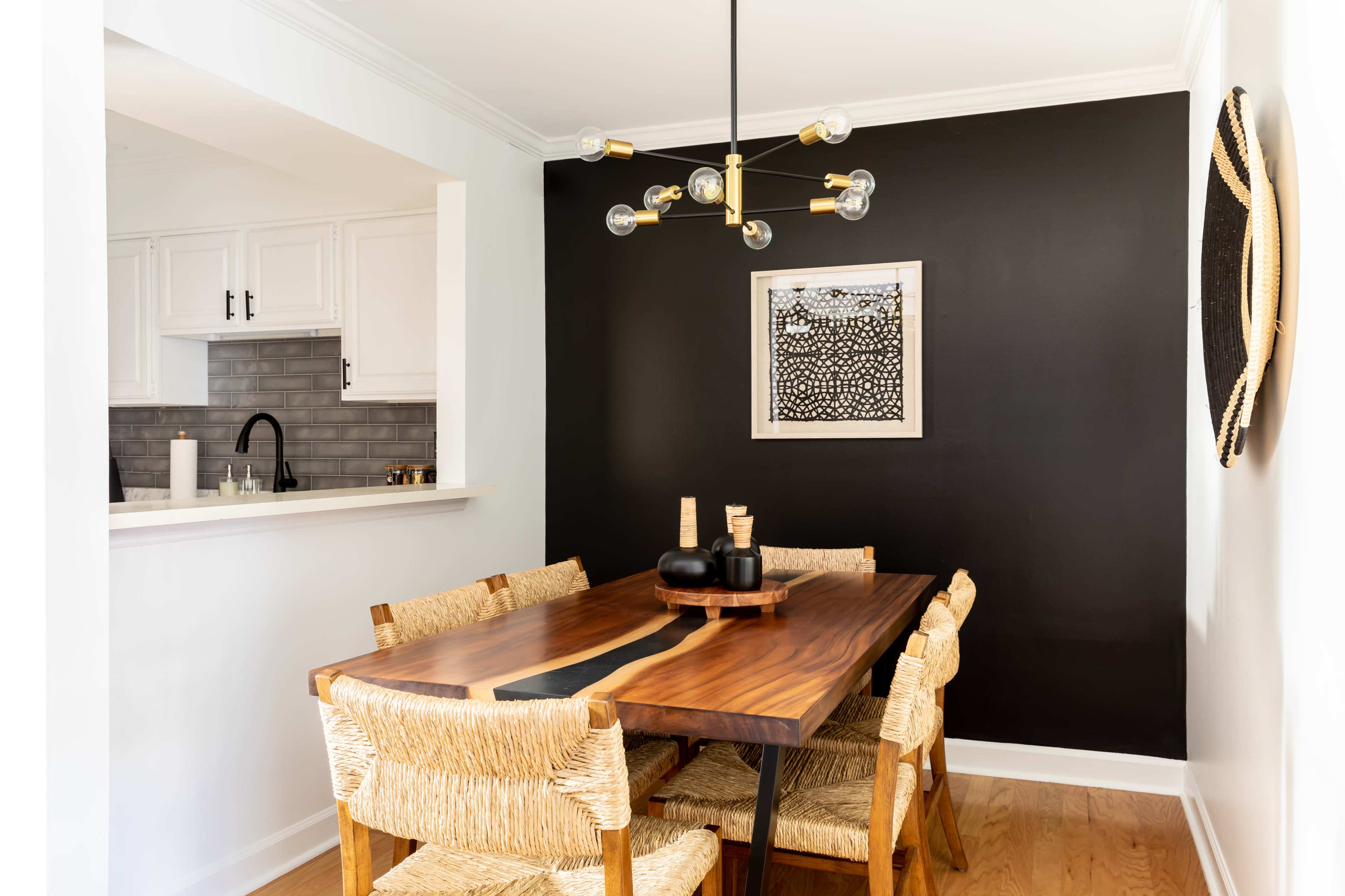 A dining area features a wooden table surrounded by woven chairs, with a black accent wall adorned by a framed artwork and a modern light fixture overhead.
