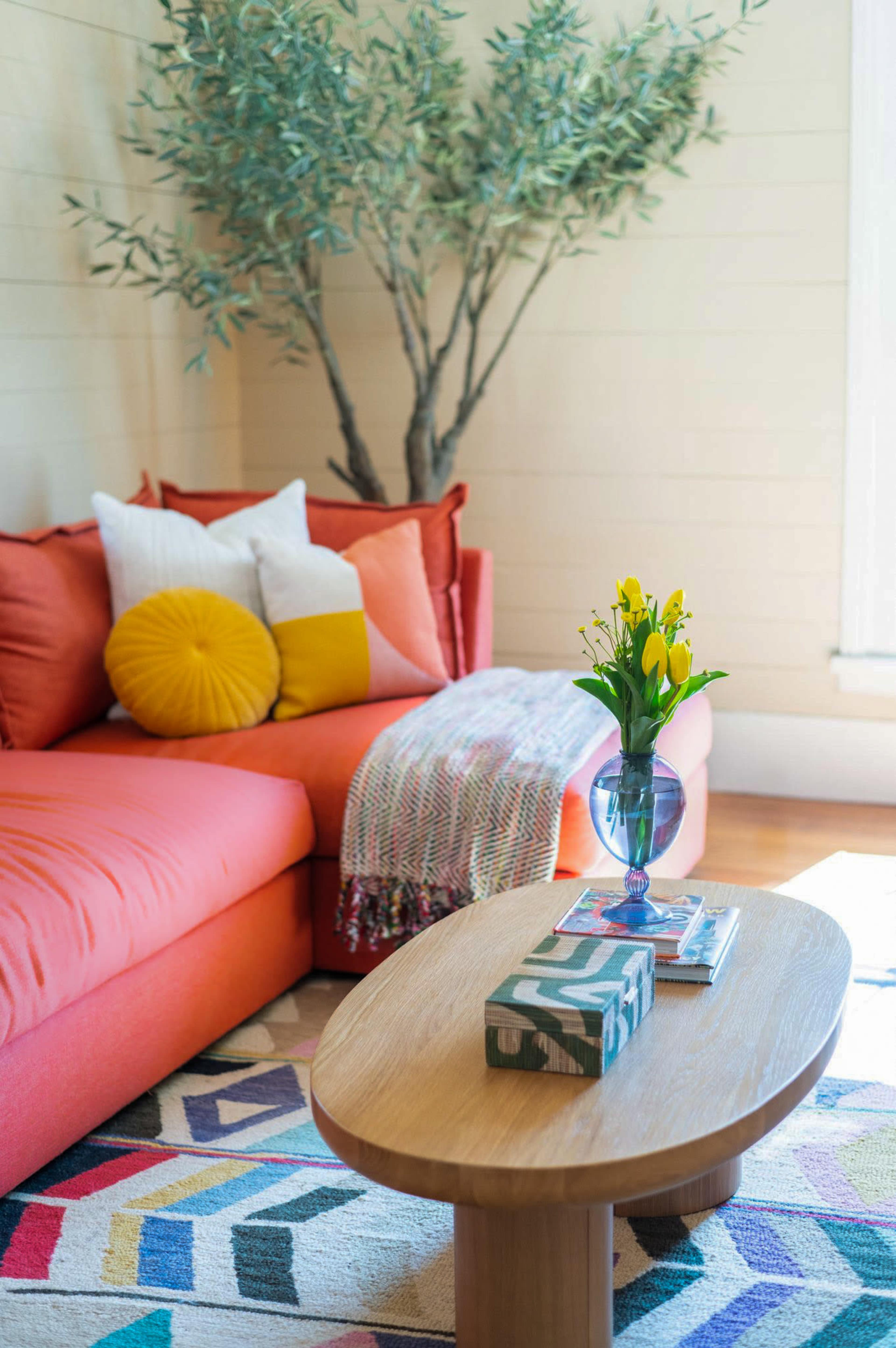 The image shows a cozy living room with an orange couch, a small wooden coffee table, a vase of yellow flowers, and a decorative throw blanket draped over the couch.