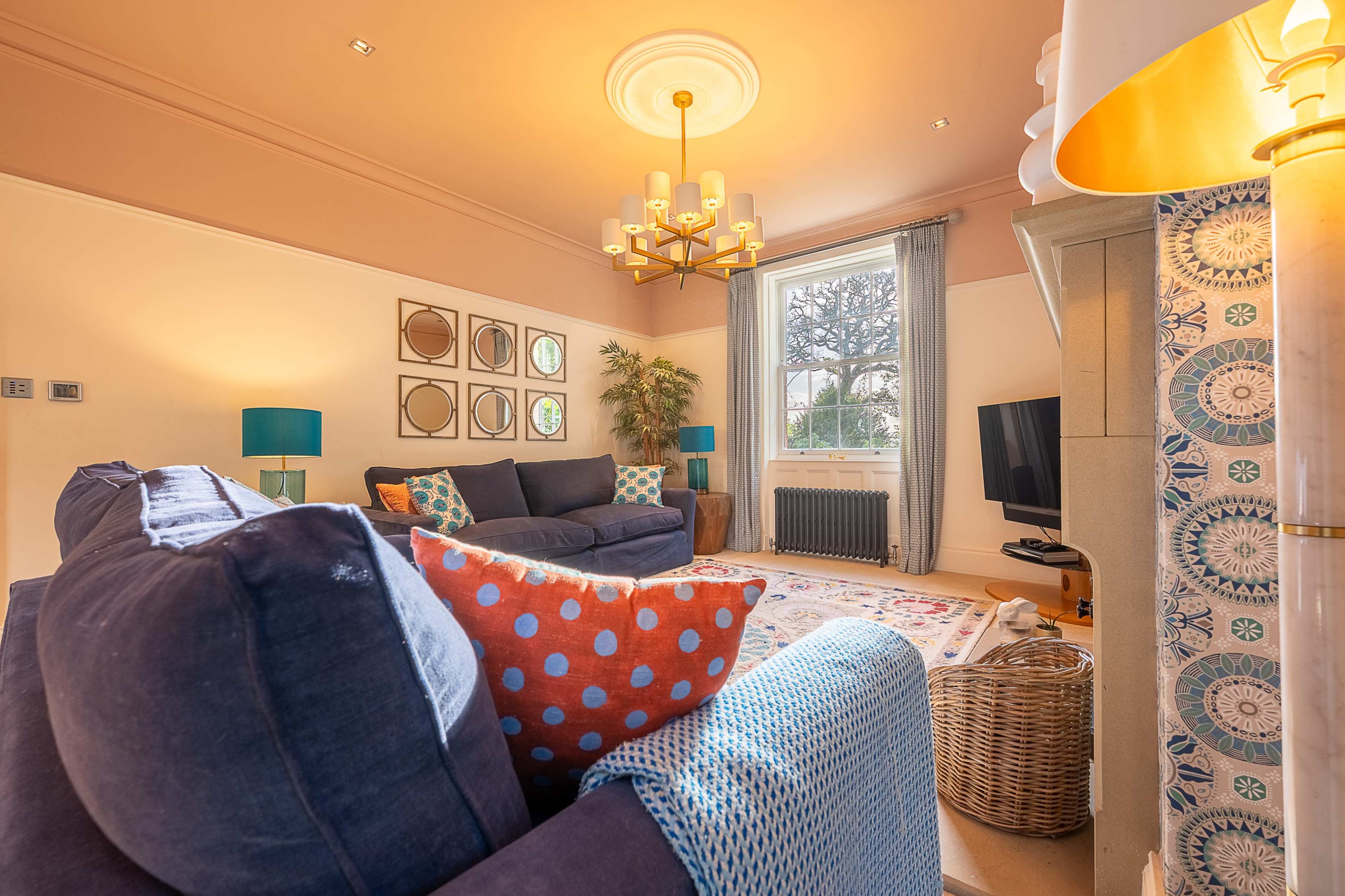 The living room features a blue sofa with patterned cushions, a chandelier, a window with a view of trees, and a television near a decorative column.