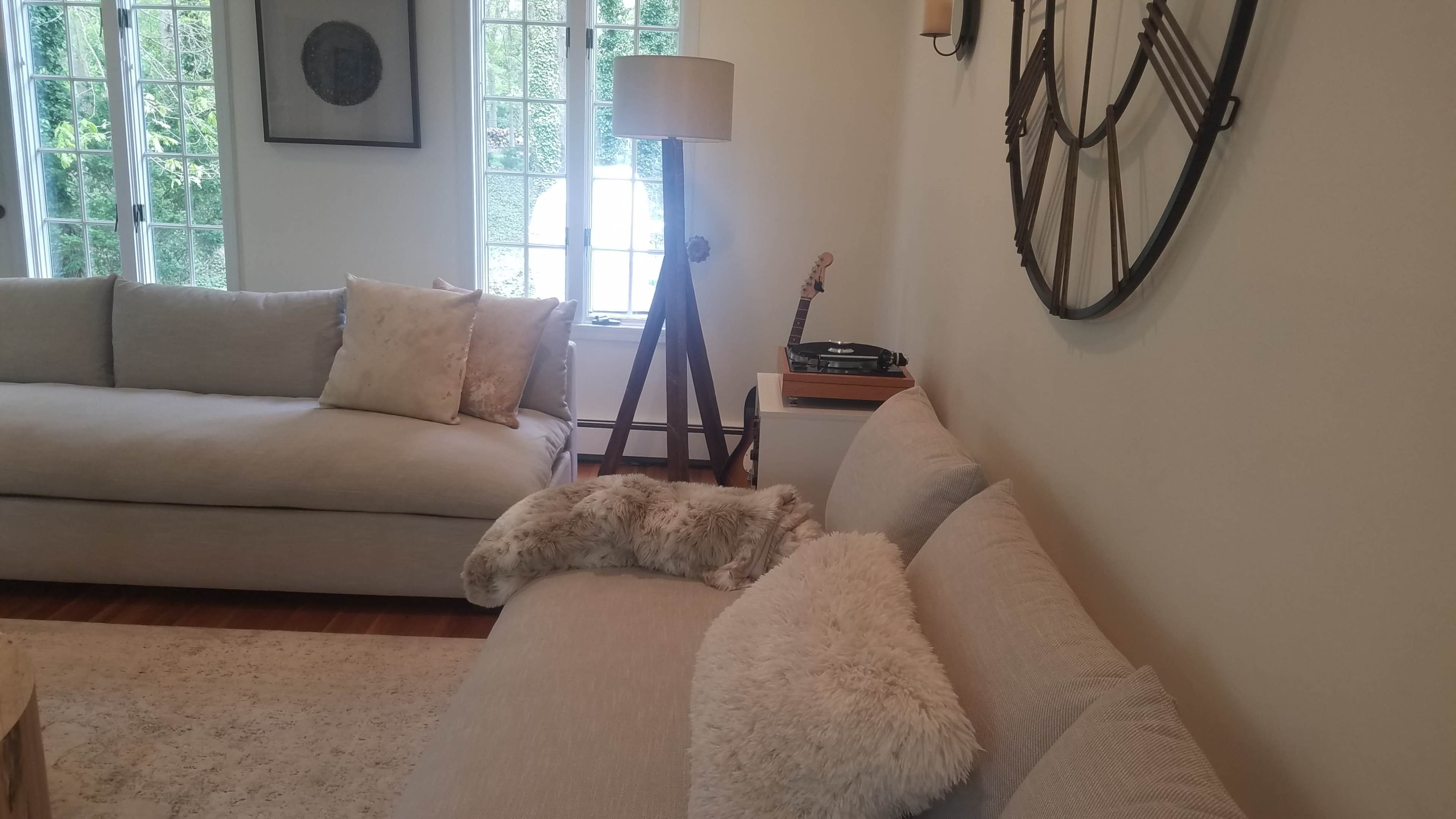 The image shows a living room featuring a light-colored sofa, a wooden lamp, and a record player next to a wall with a large clock.
