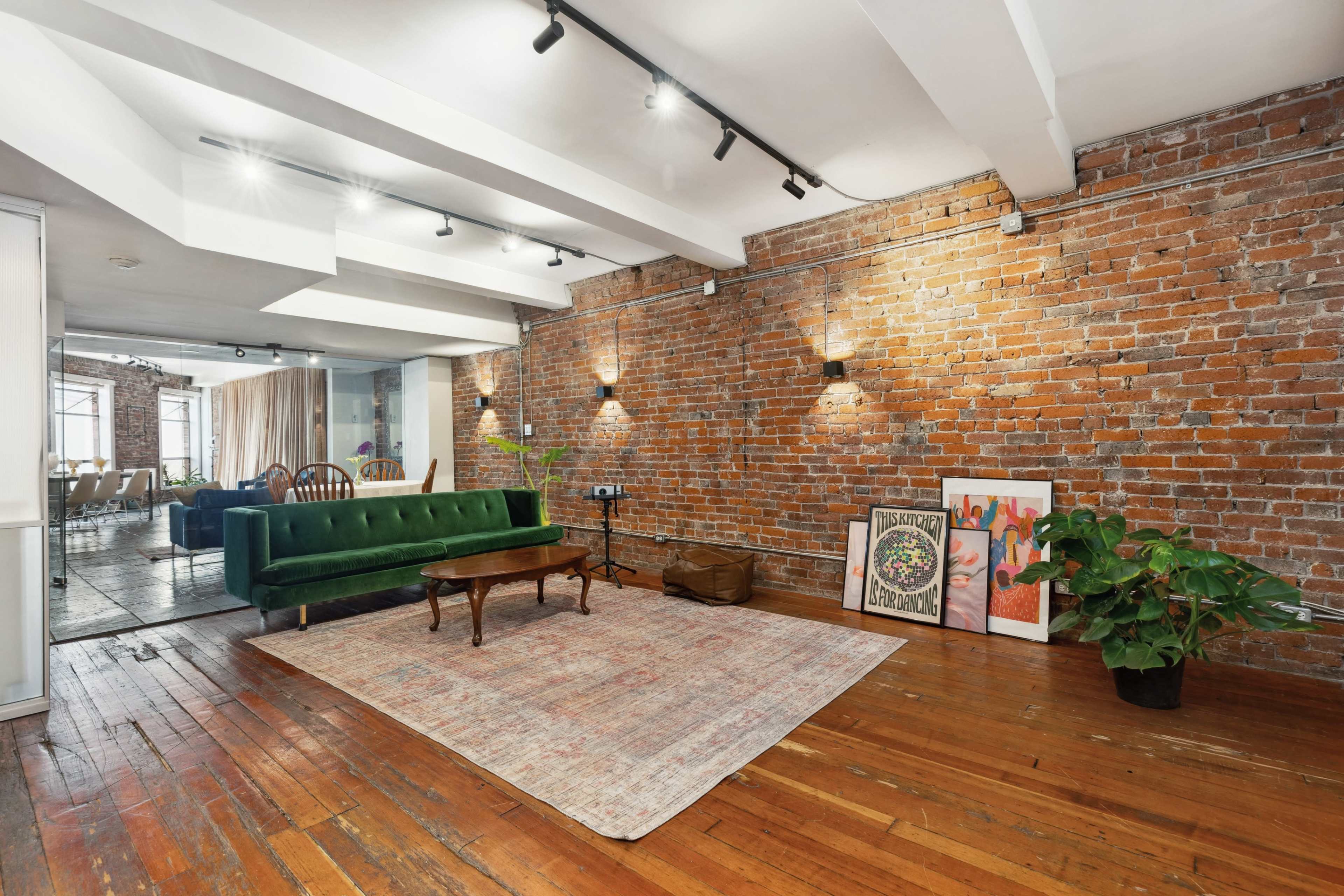 A spacious room with exposed brick walls, wooden floors, a green velvet sofa, a wooden coffee table, and decorative artwork on the floor.