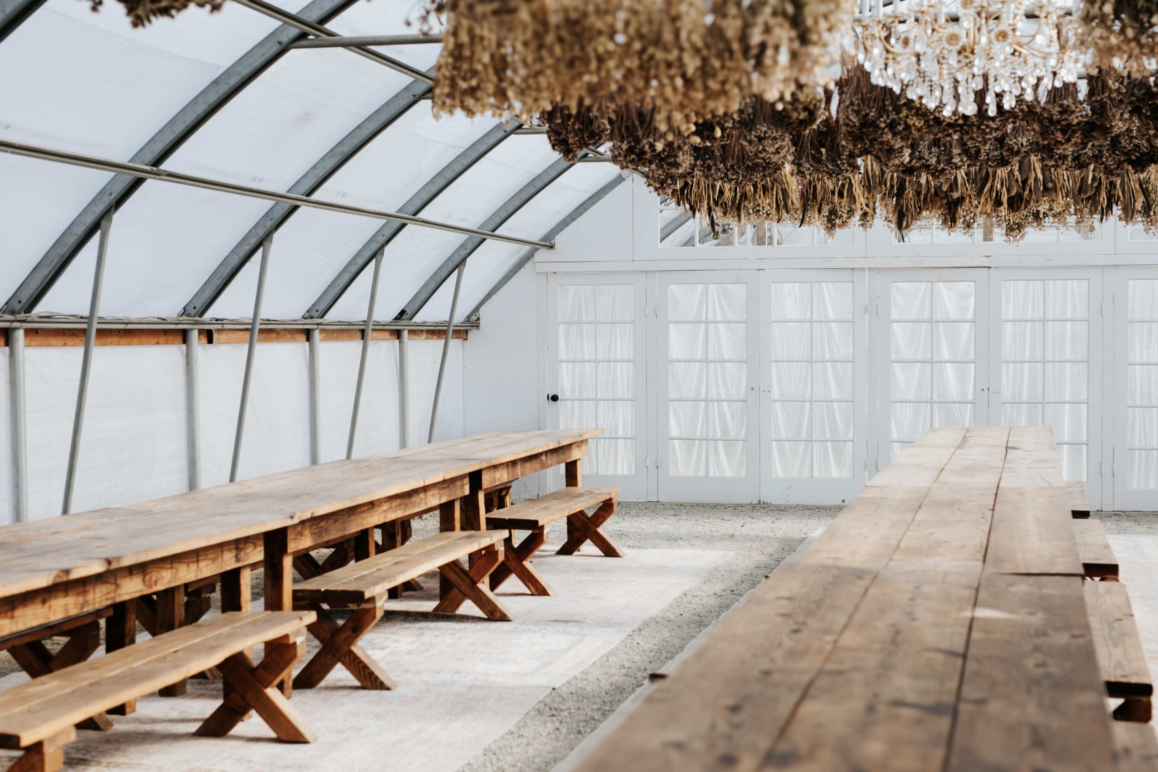 A spacious indoor greenhouse features long wooden tables and benches beneath a decorative arrangement of dried plants and a chandelier.