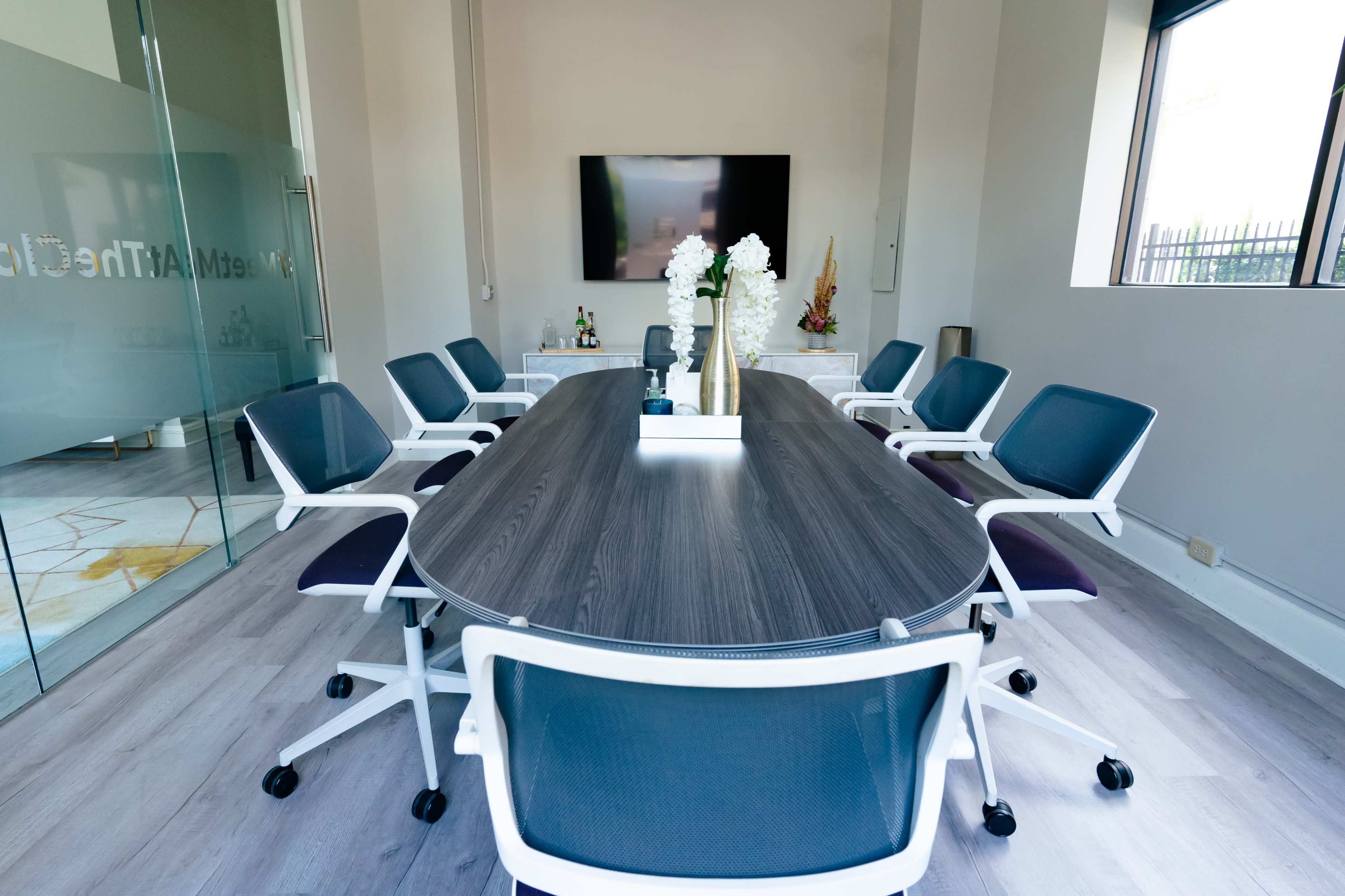 A modern conference room features a large oval table surrounded by white and gray rolling chairs, with a television mounted on the wall and a decorative centerpiece.