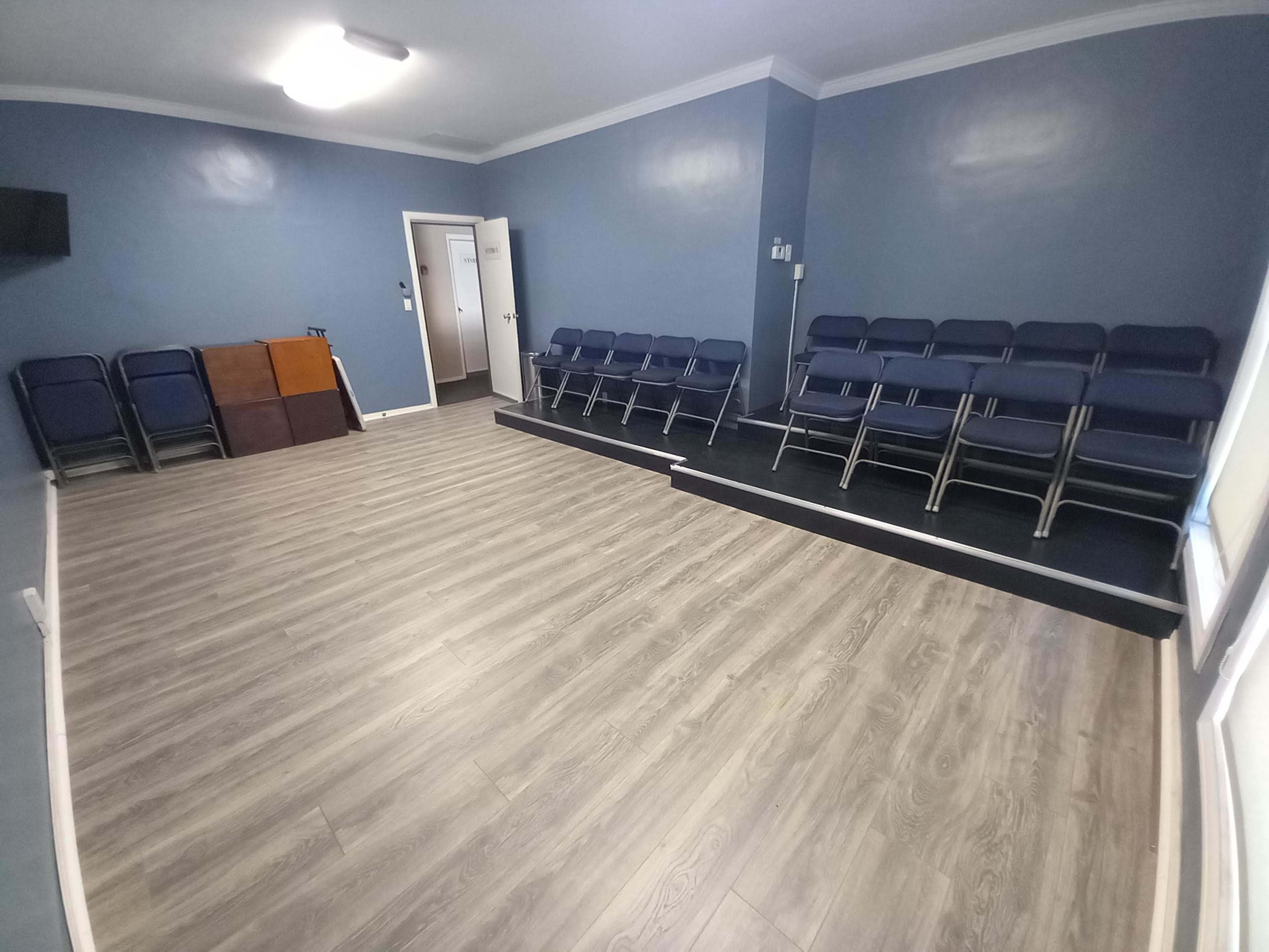 A simple room with blue walls, a wooden floor, a set of stacked chairs on a platform, and a doorway leading to another space.