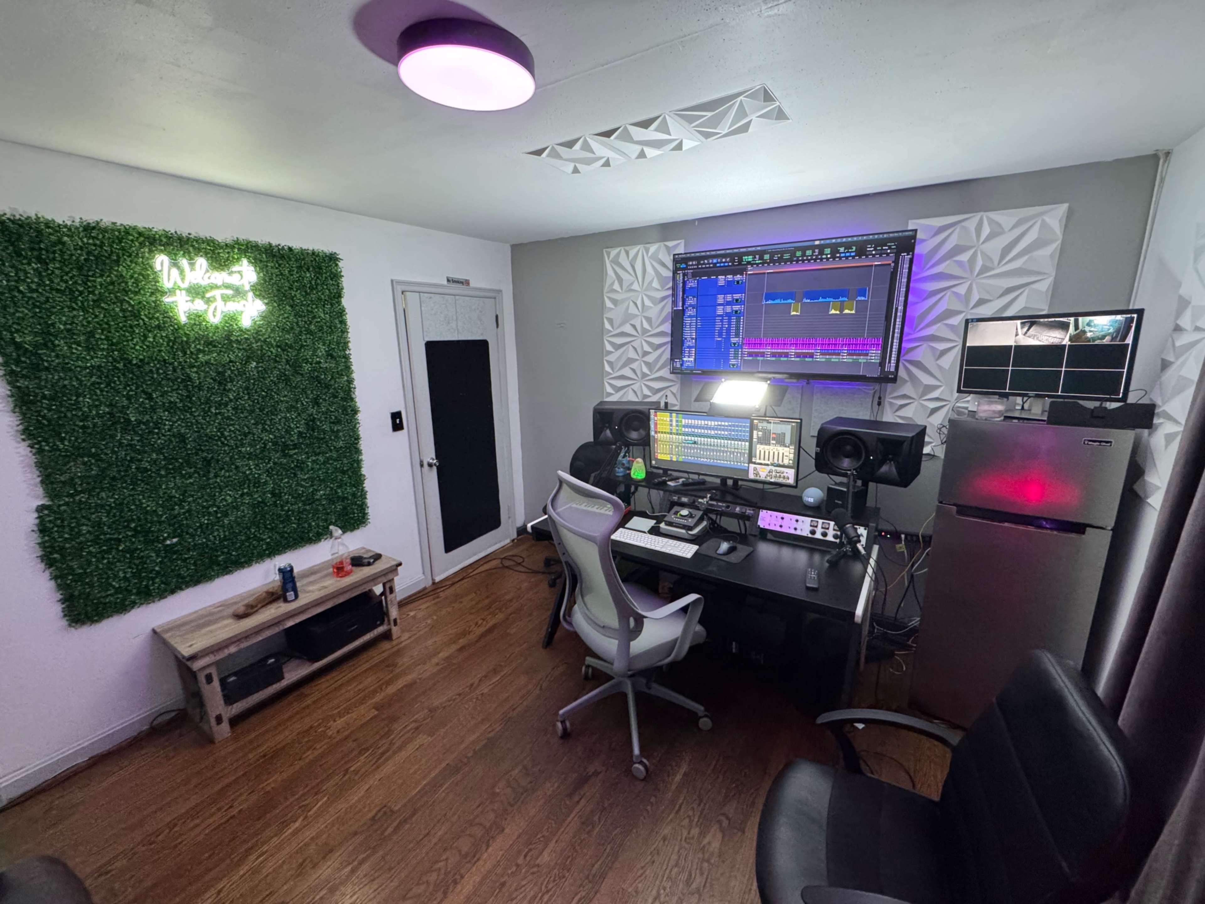 The image shows a modern music production studio featuring a desk with multiple monitors, a sound system, and decorative greenery on the wall.