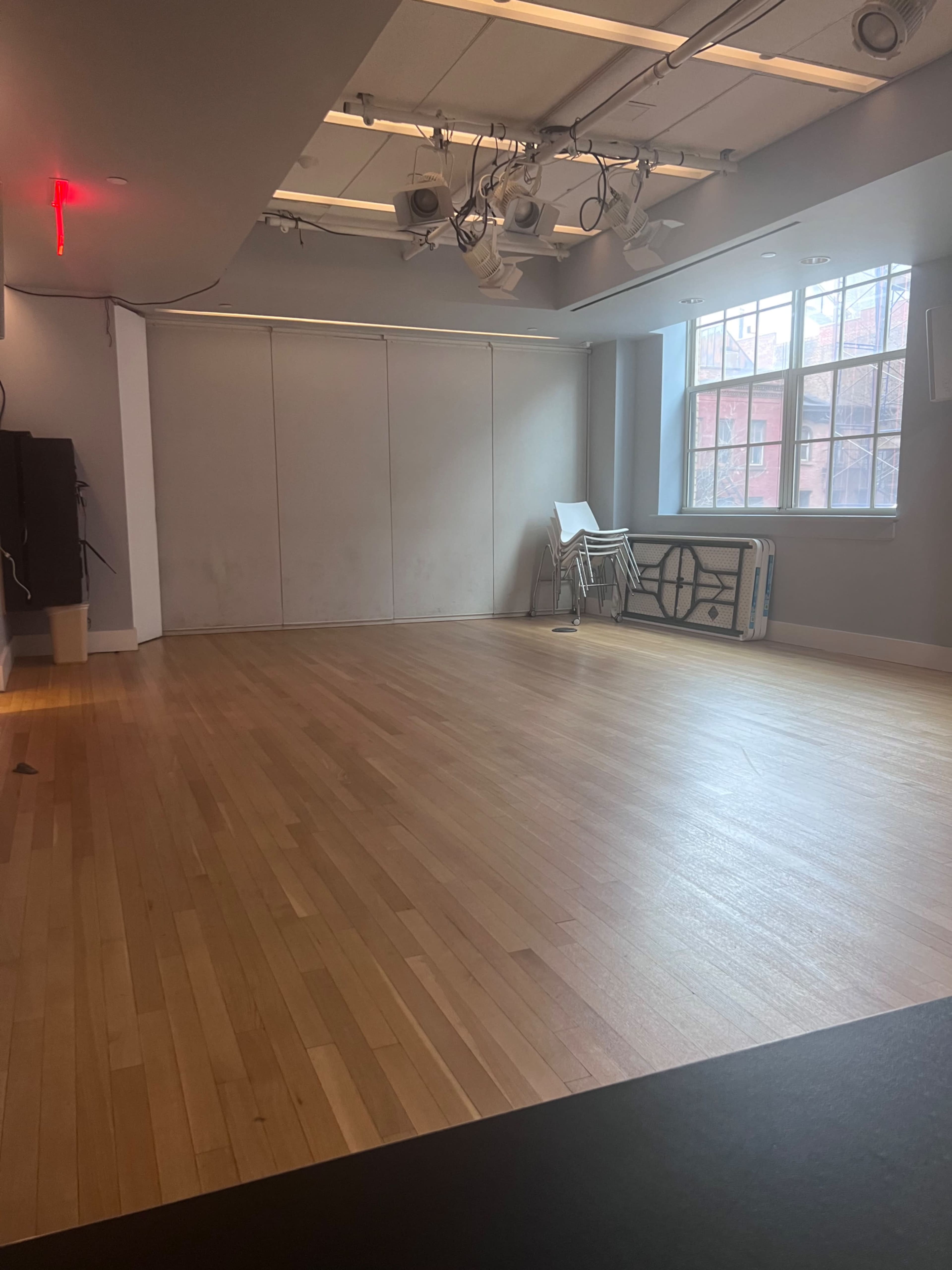 The image shows a spacious, empty room with hardwood flooring, large windows, and a stack of folding chairs against one wall.